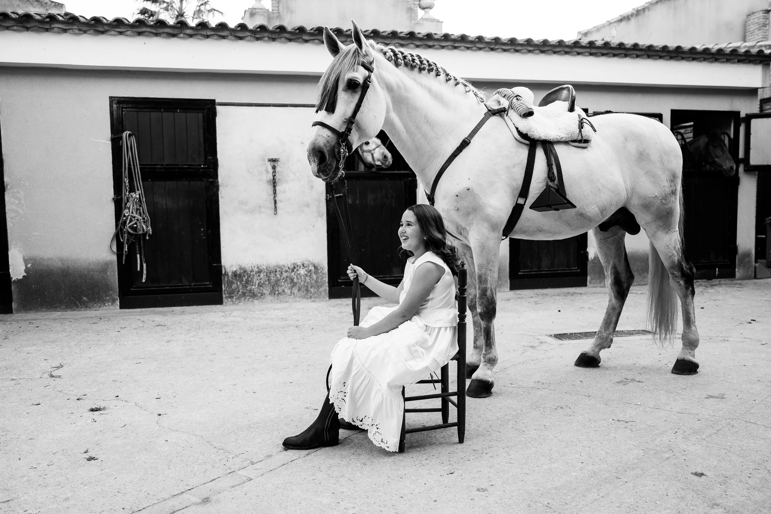 Una niña vestida con un vestido blanco, sentada en una silla, sosteniendo las riendas de un caballo de color blanco con crines trenzadas y equipada con arnés, en un patio con paredes blancas y rejas negras.