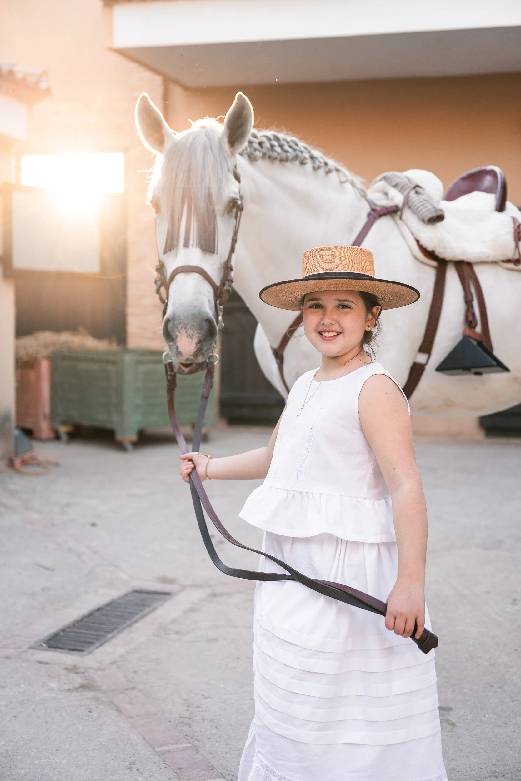 Niña sonriendo, sosteniendo la correa de un caballo blanco en un lugar cubierto de luz cálida.
