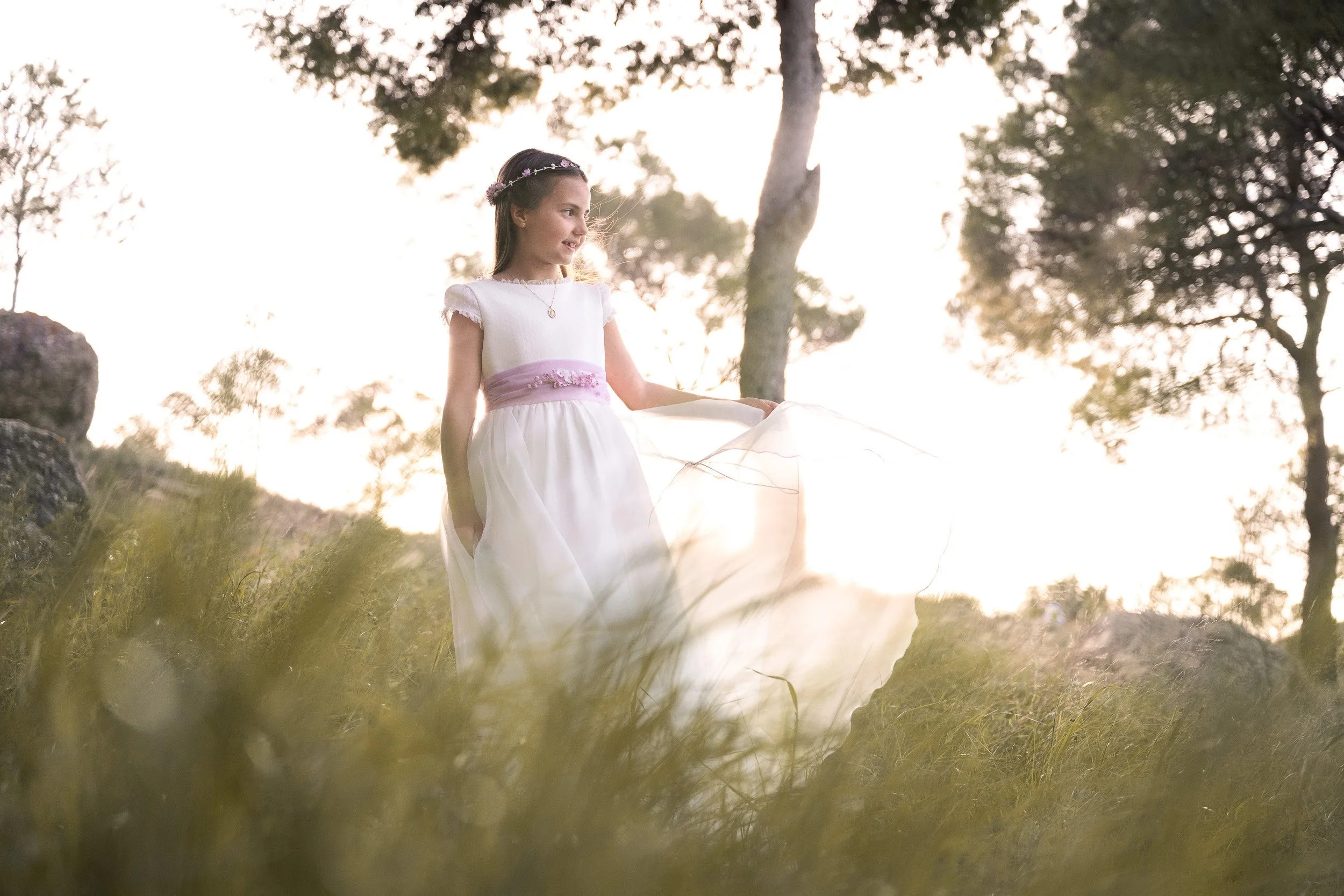 Una niña con vestido de comunión y corona de flores en un entorno natural con árboles y césped durante el atardecer.