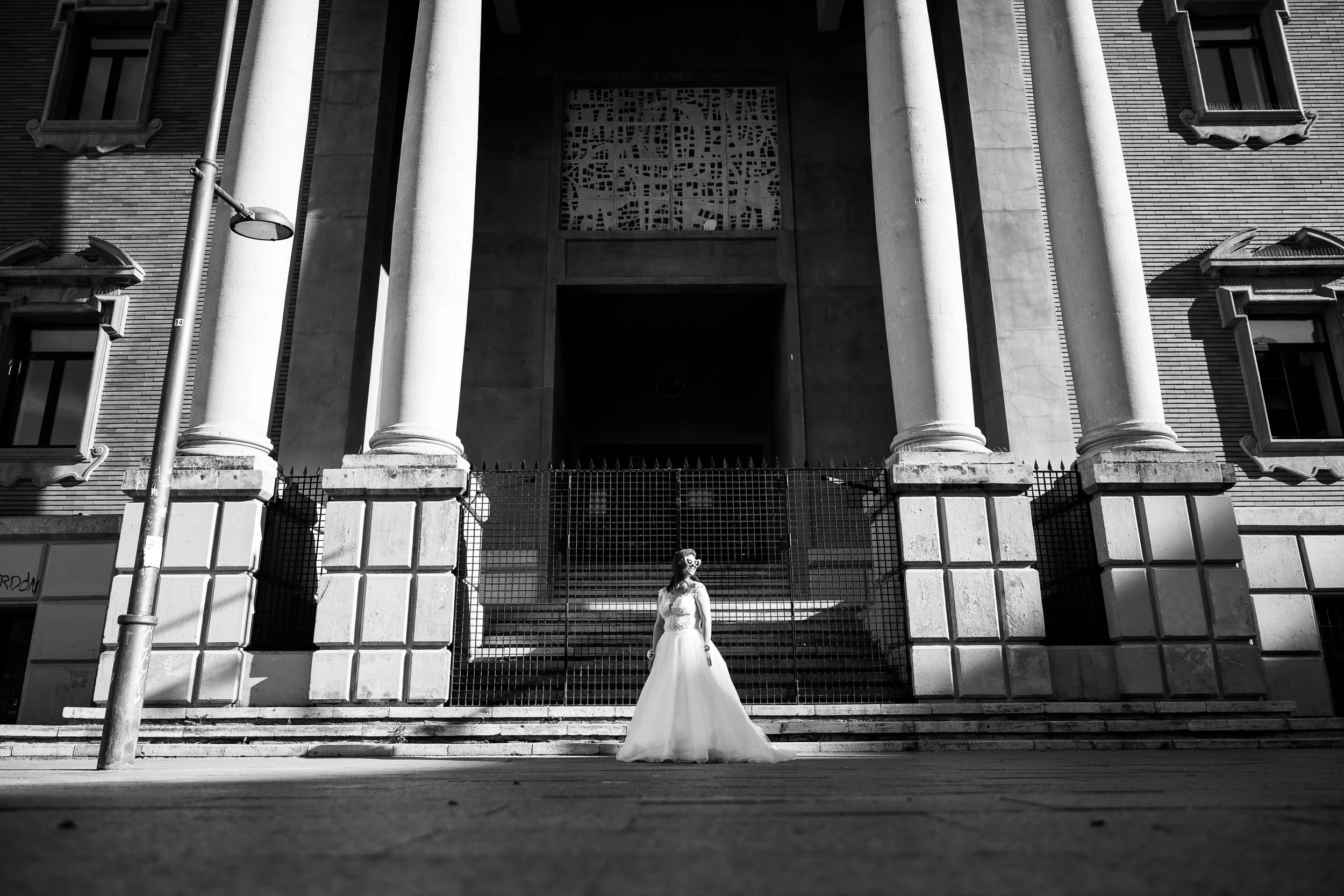 Mujer con vestido de novia de pie frente a un edificio grande con columnas y ventanas en blanco y negro.