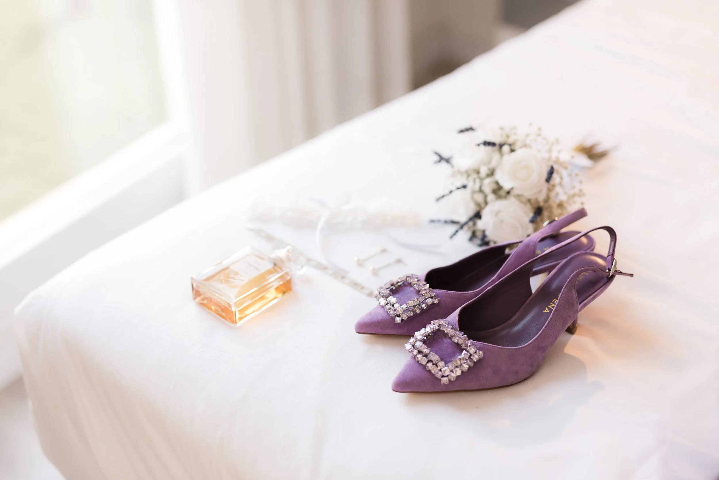 Zapatos de tacón morados con adornos de cristales, un perfume en frasco rectangular y una pulsera con cristales sobre una cama blanca