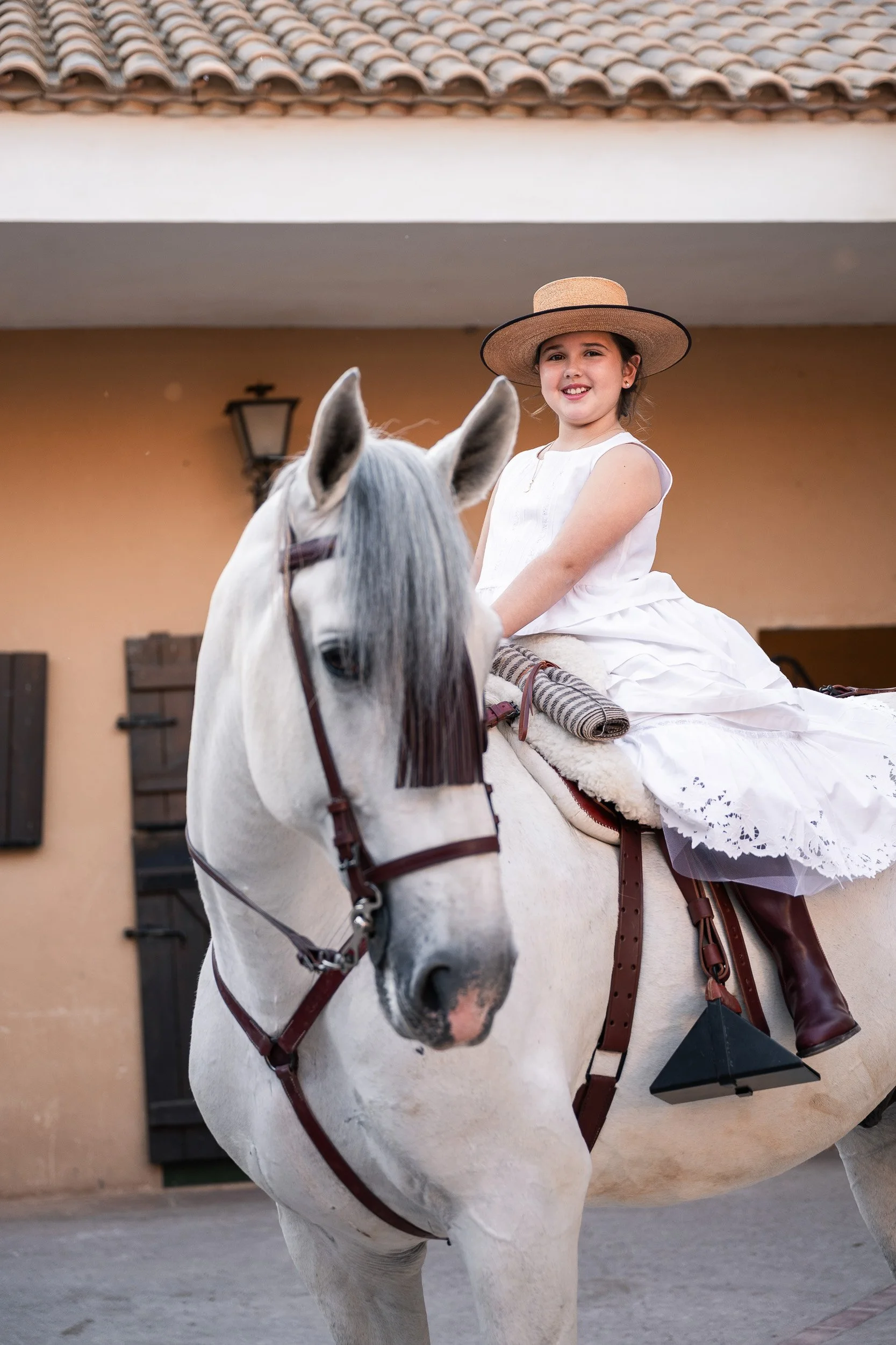 Niña joven montada en caballo, viste vestido blanco y sombrero de paja, en un entorno exterior con pared beige y farol antiguo.