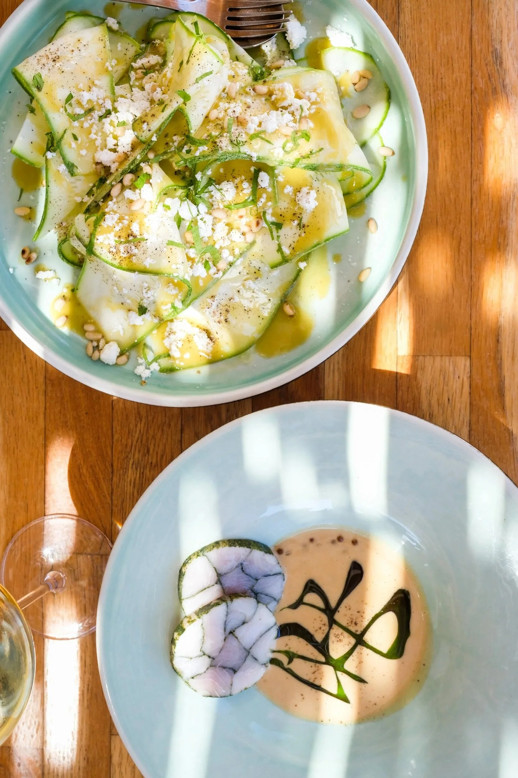 Close-up of two bowls, placed on a wooden table. One bowl contains a fish mosaic of fresh fish hand rolled in a sauce. The other dish is a zucchini salad, thinly sliced with local cheese, pine nuts and garnished with herbs