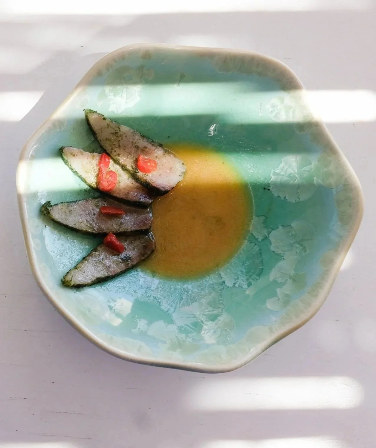 An aqua green ceramic bowl with semi cured amberjack in a kombucha butter sauce with small pieces of dried tomato