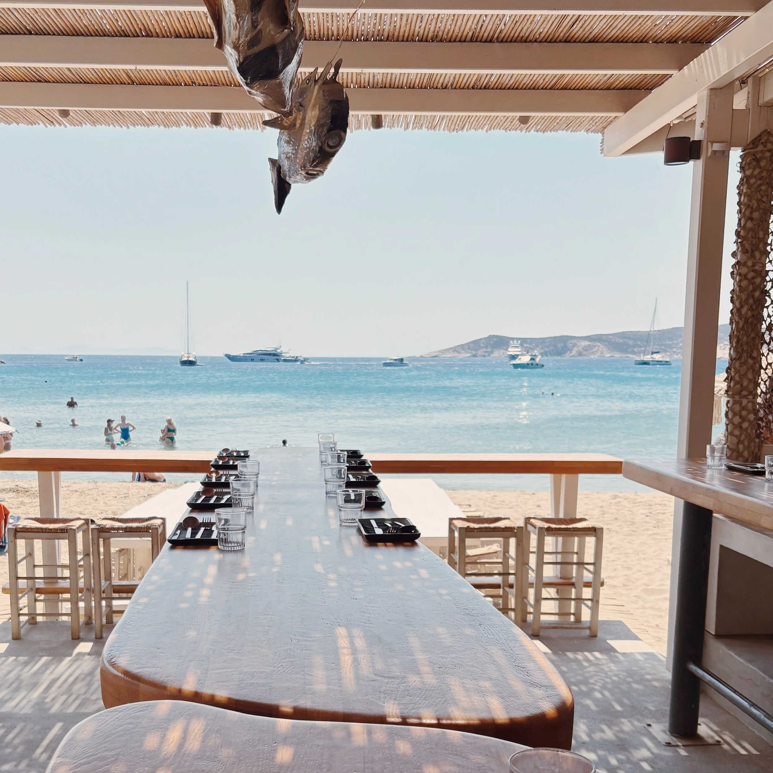 View from inside a beachside restaurant / bar, showing a beach with boats in the water, and another island in the distance. The foreground features a long wooden dining table, high stools, fish pendant lights and part of the restaurant's structure.