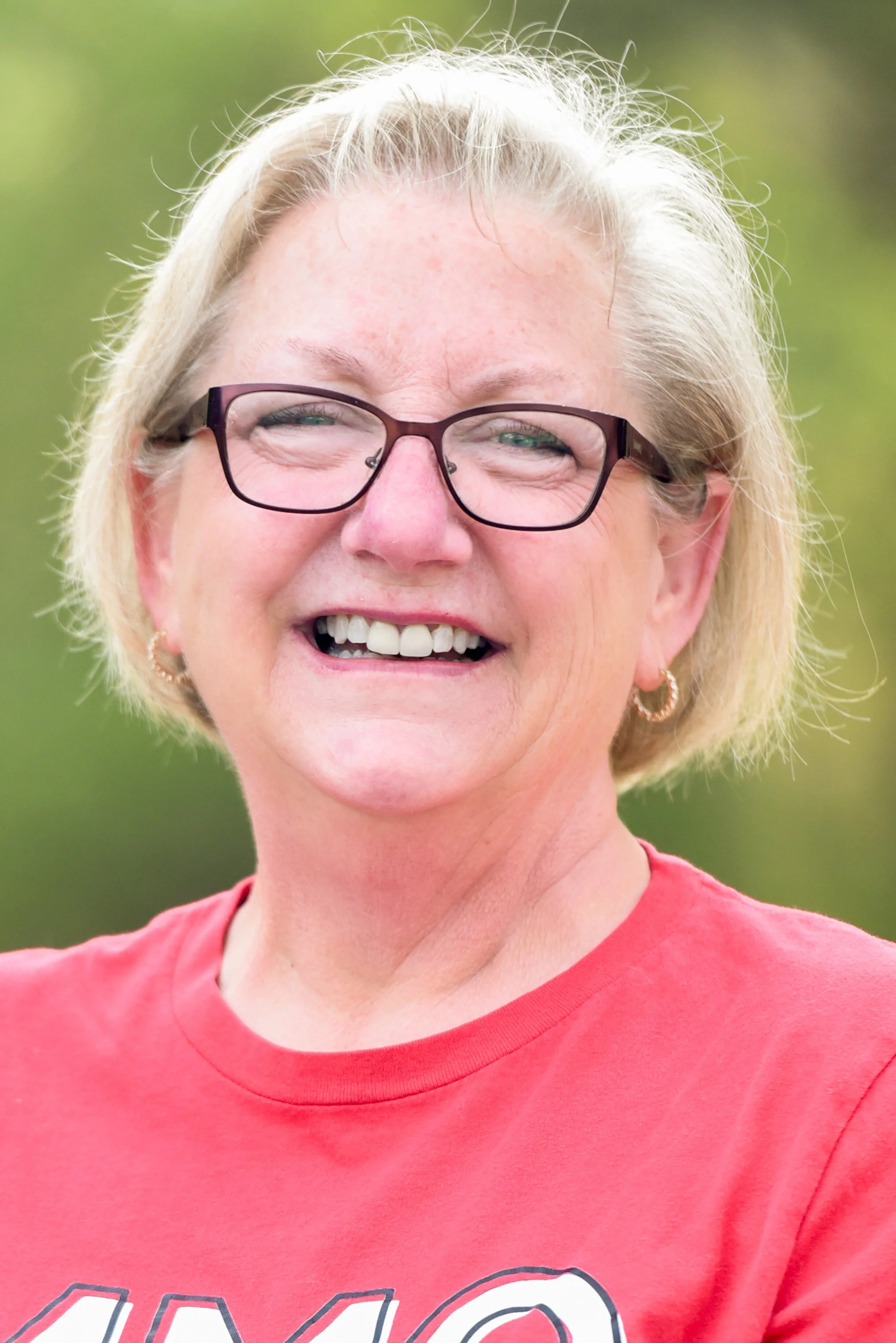 A woman with short blonde hair, wearing glasses and a red shirt, smiling outdoors with a blurred green background.