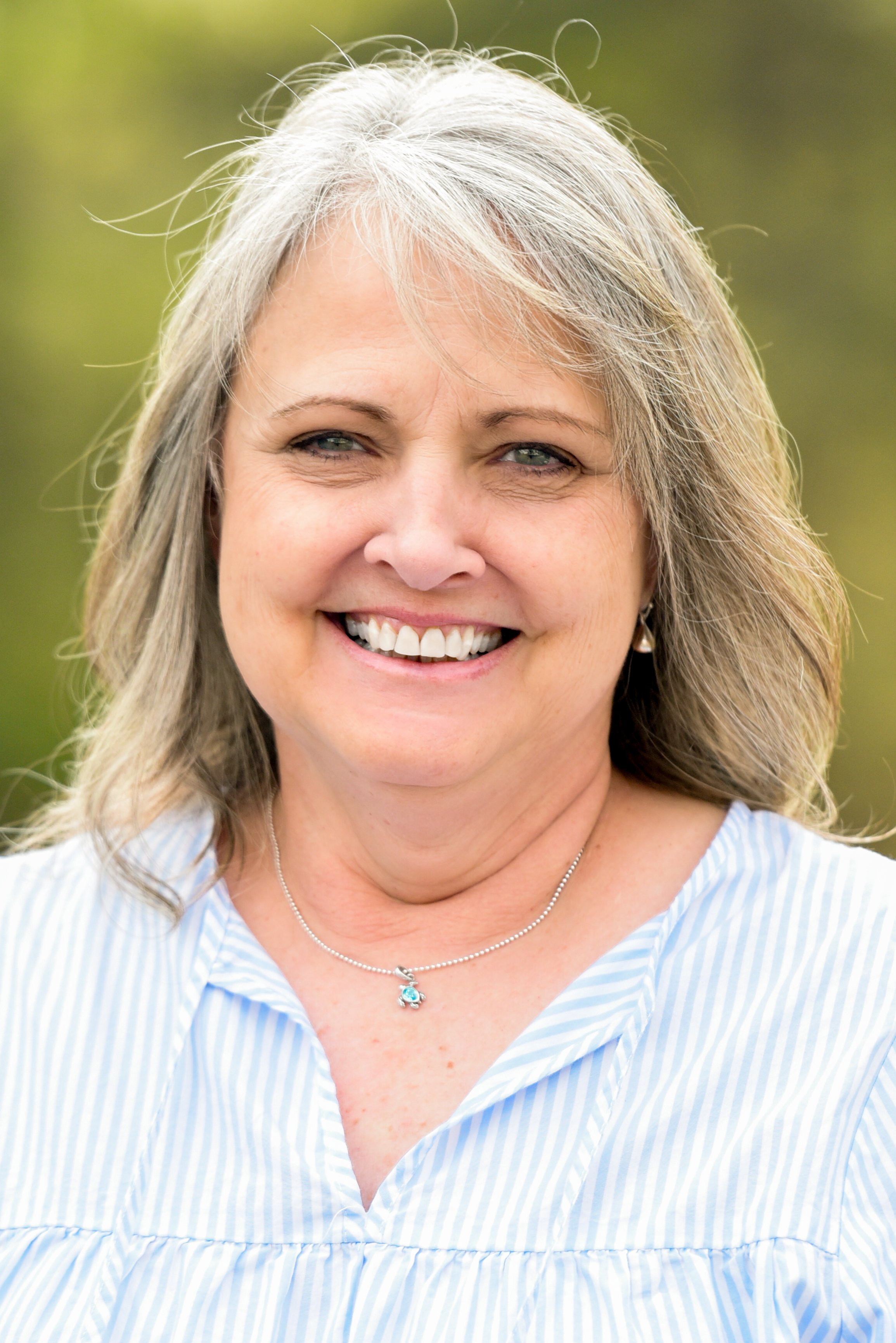 A woman with long gray hair smiling outdoors, wearing a light blue and white striped blouse and a silver necklace with a blue pendant.