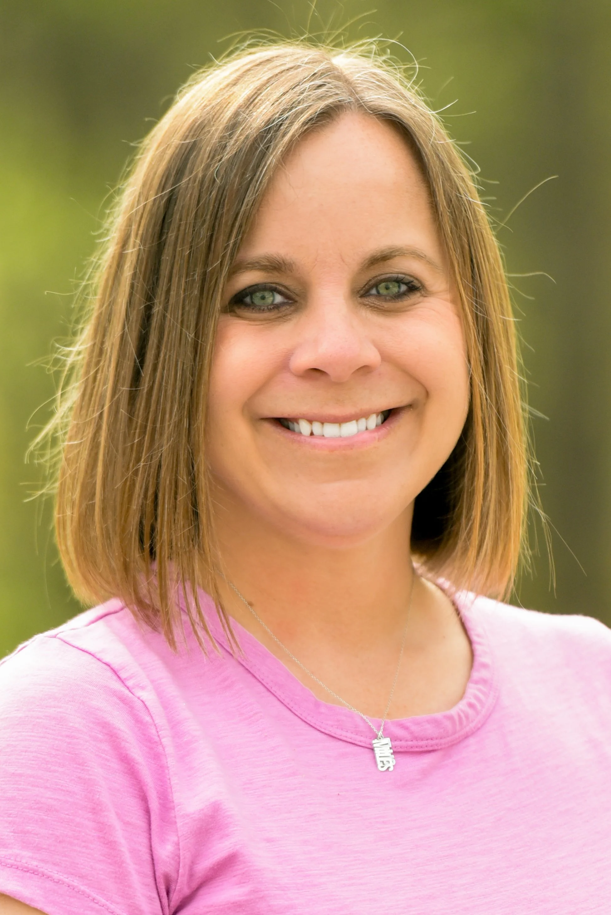 Close-up of a woman smiling outdoors, wearing a pink shirt and a silver necklace with a 'Smile' pendant.