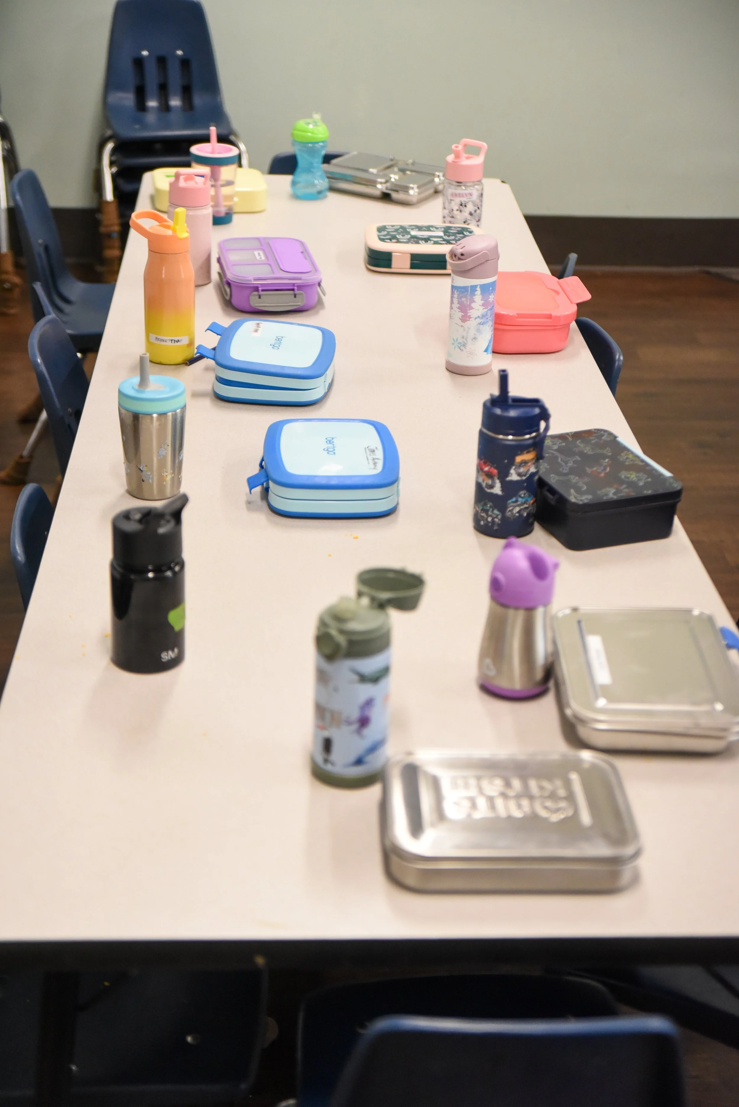A long white table filled with colorful reusable water bottles, stainless steel containers, and lunch boxes with blue, pink, purple, and green lids in a room with dark wood flooring and dark chairs.