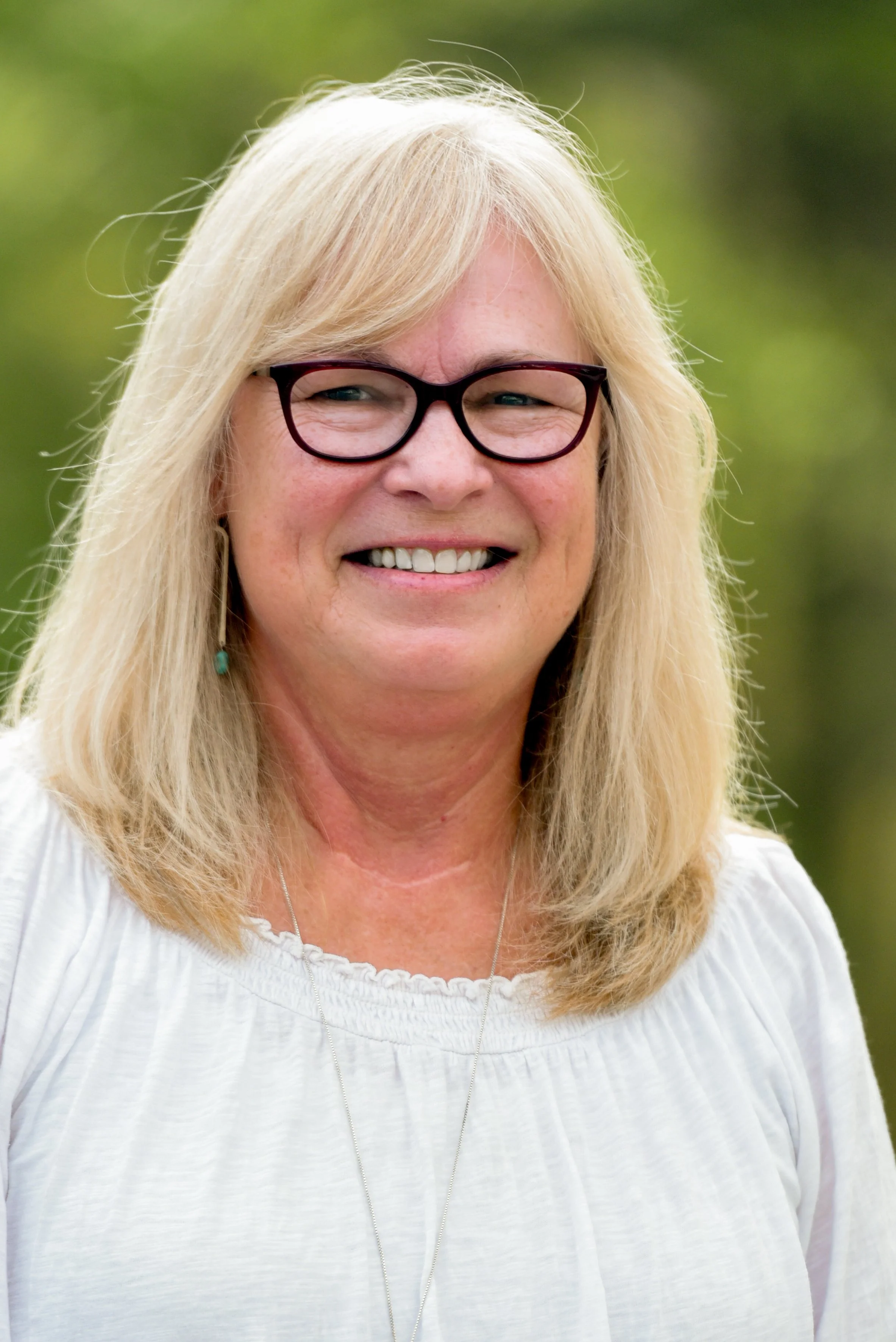 Close-up of a smiling woman with blonde hair wearing glasses and a white top, outdoors with green out-of-focus background.