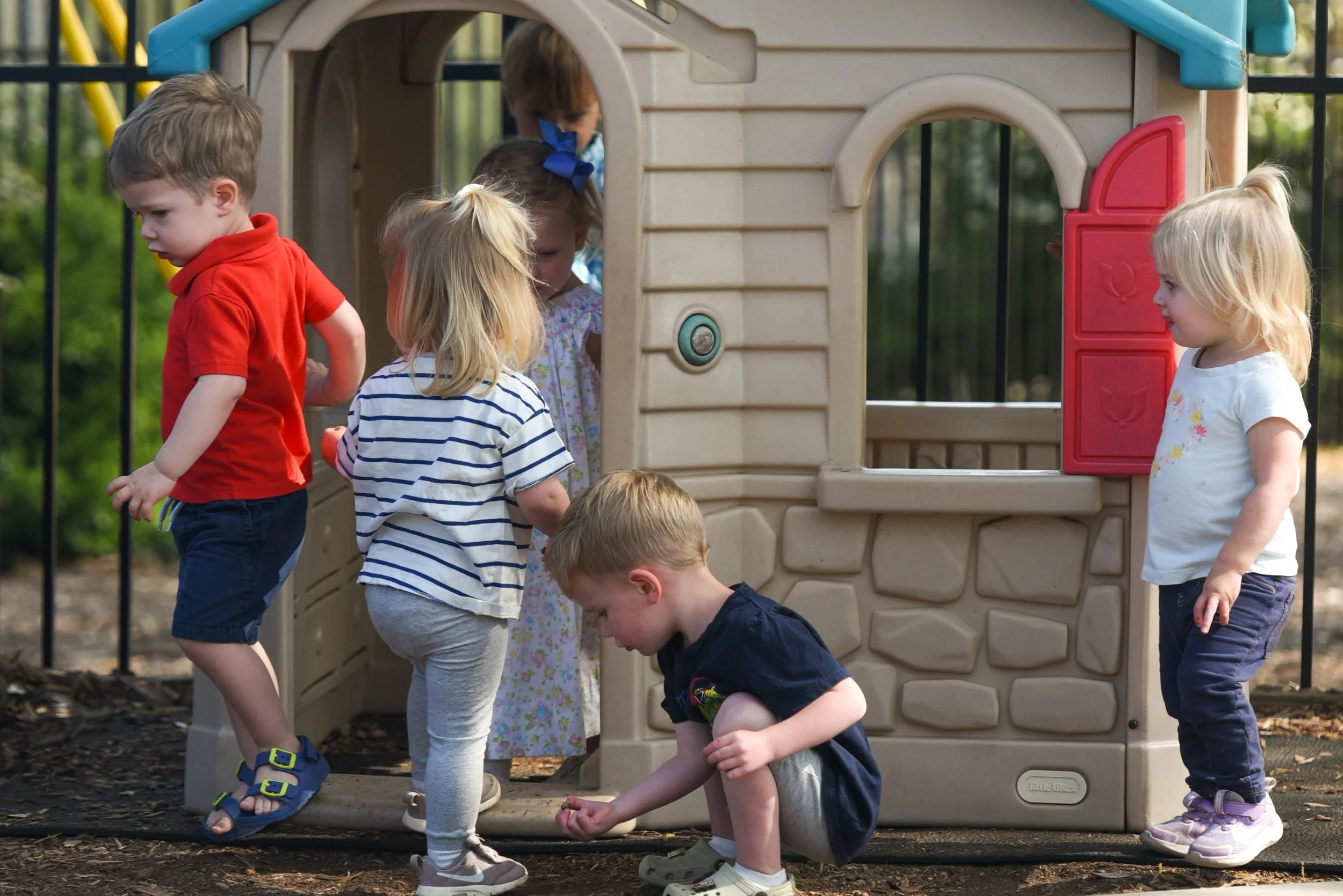 Children playing outside by a small plastic playhouse at a playground.