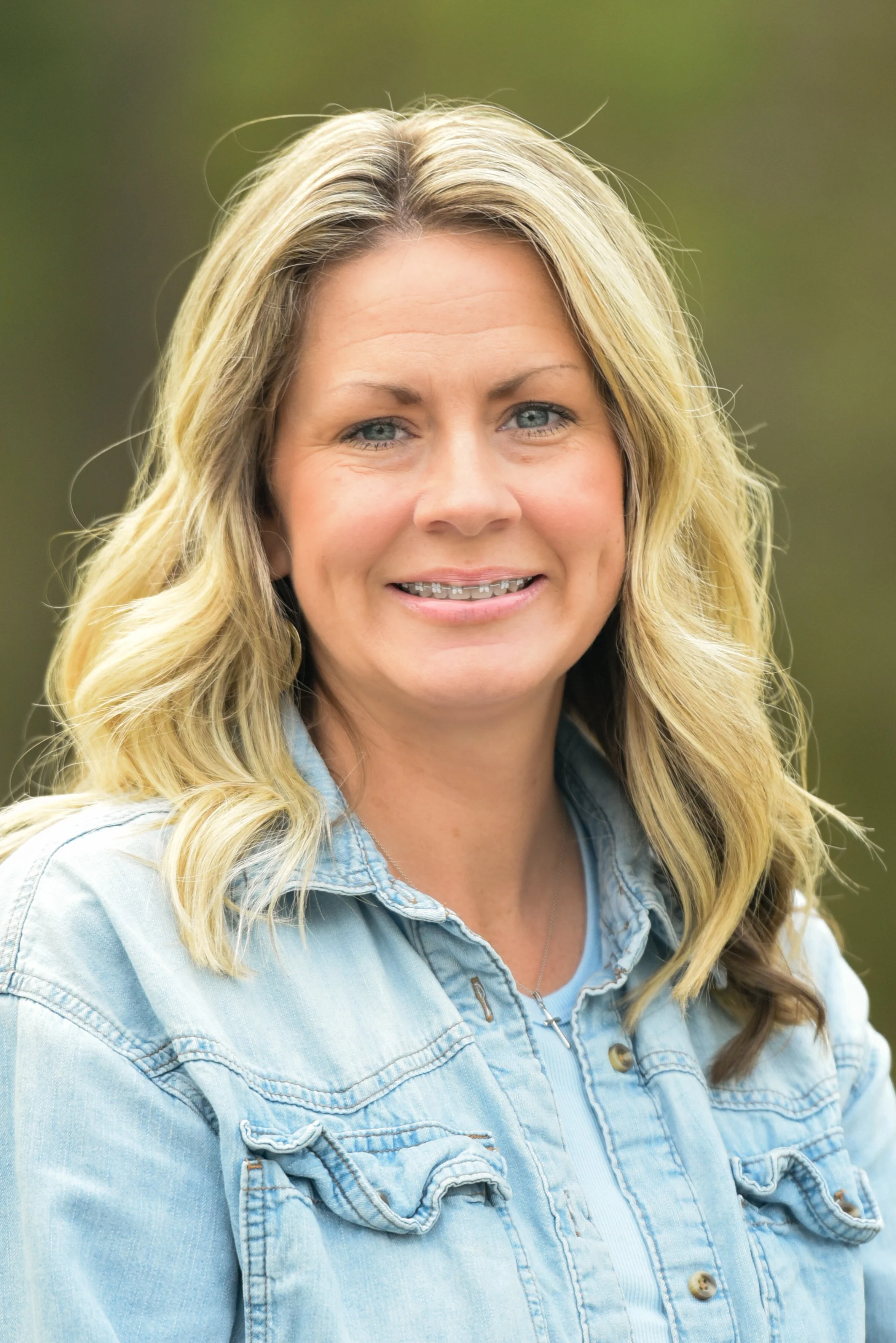 Close-up portrait of a woman with blonde wavy hair wearing a light blue denim jacket, smiling outdoors with a blurred green background.
