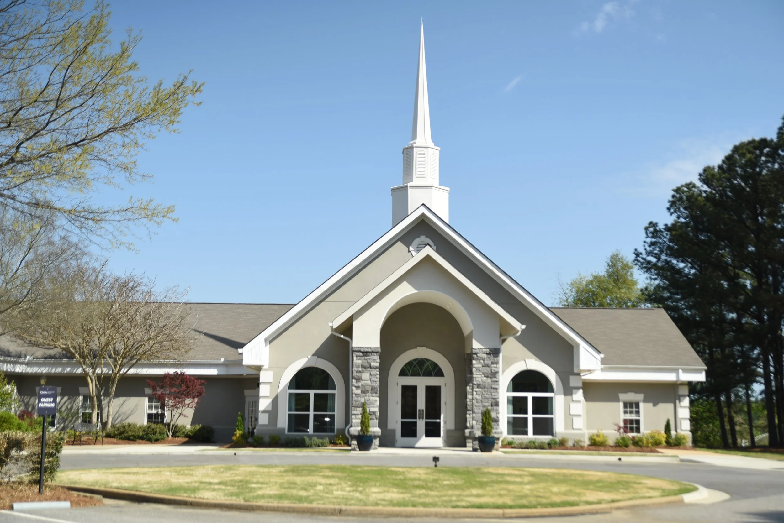 Front view of a white church building with a tall steeple, arched windows, and a landscaped yard with trees and shrubs.