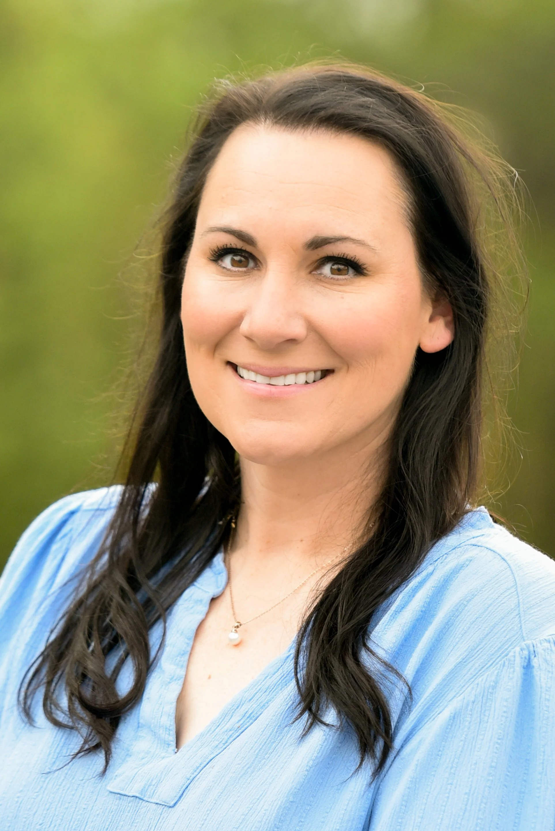 Close-up portrait of a woman with long dark hair, wearing a light blue shirt and a pearl necklace, outdoors with a blurred natural green background.