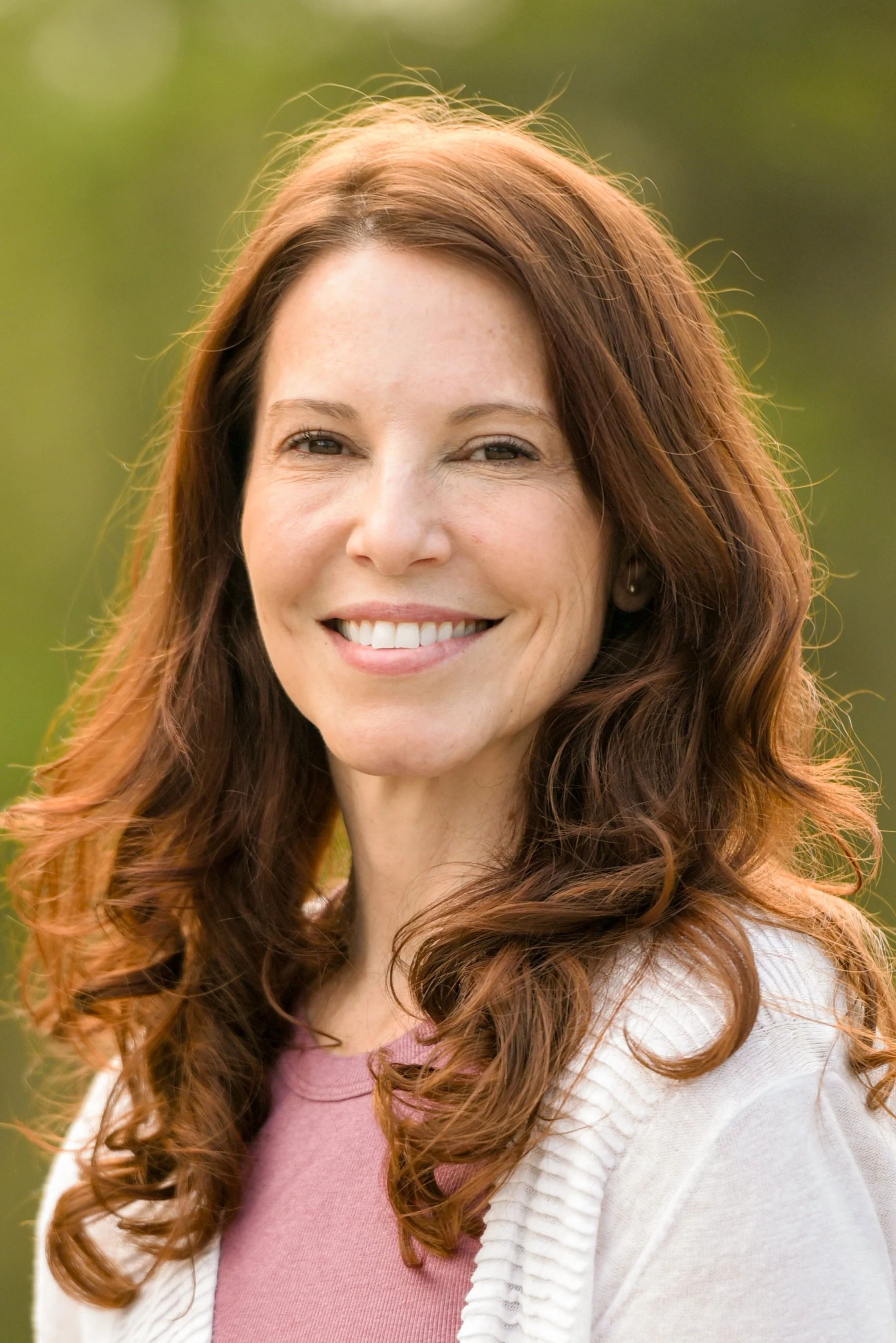 A woman with long, curly red hair smiling outdoors, wearing a pink shirt and cream-colored cardigan.