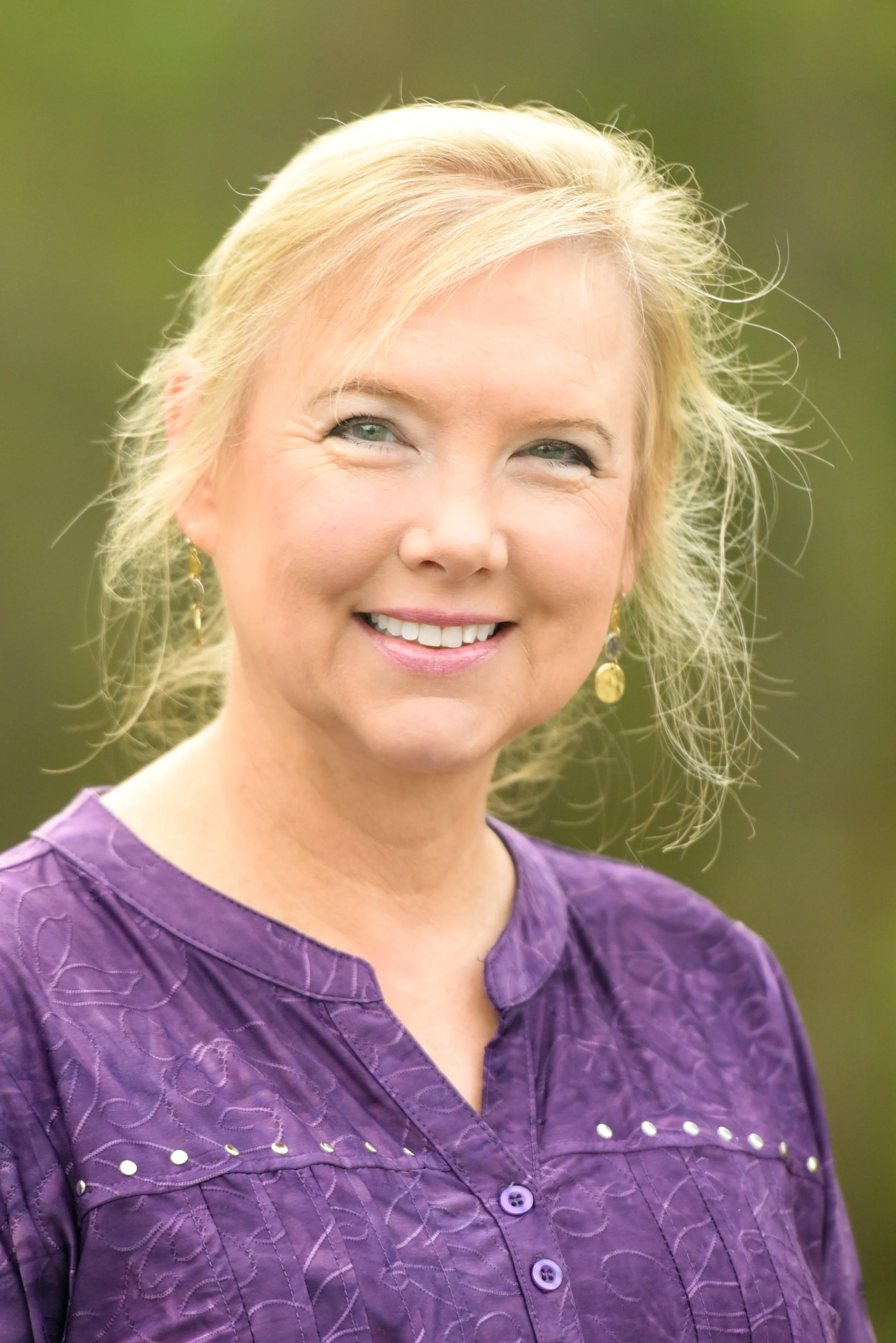 A woman with blonde hair in a loose updo, smiling, wearing a purple embroidered blouse with buttons and drop earrings, outdoors with a blurred green background.