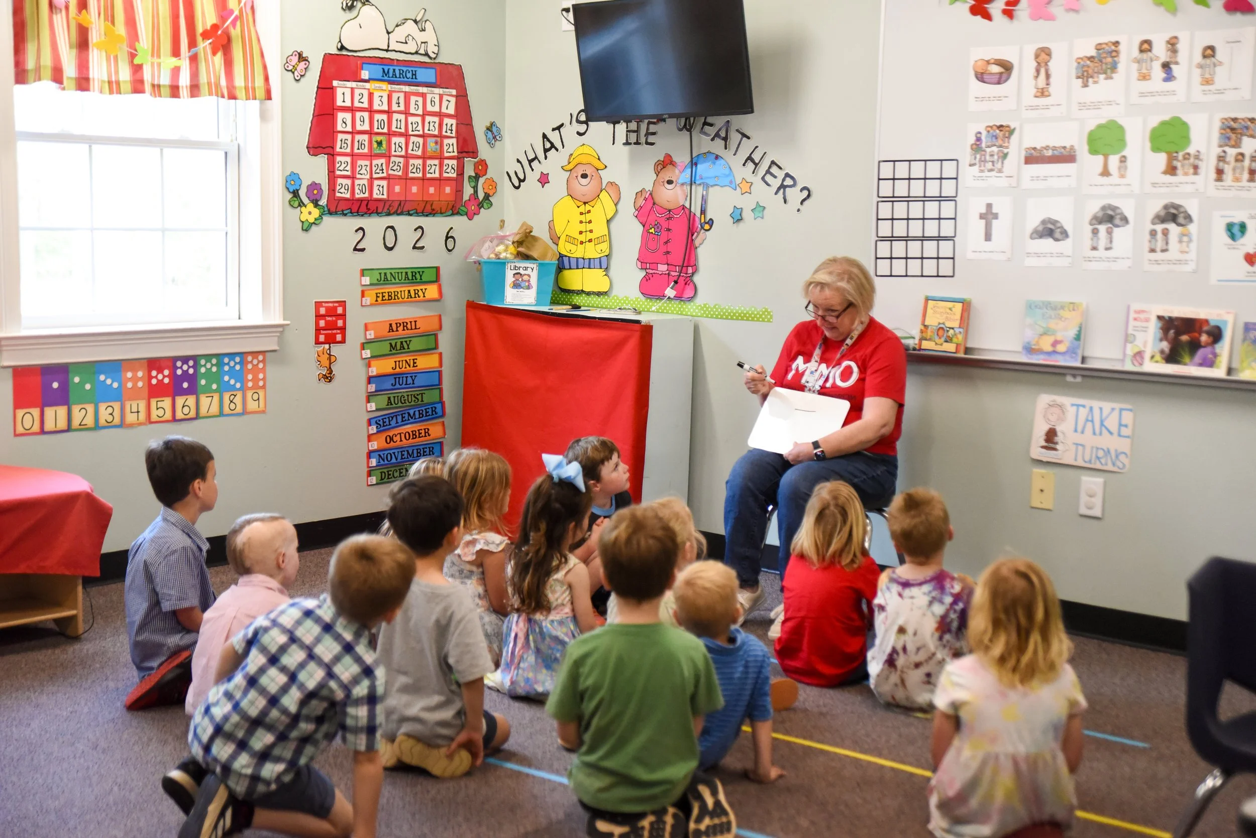 A classroom with young children sitting on the floor and a woman reading to them. The classroom is decorated with colorful educational posters, a calendar, and cutouts. A large TV is mounted on the wall.