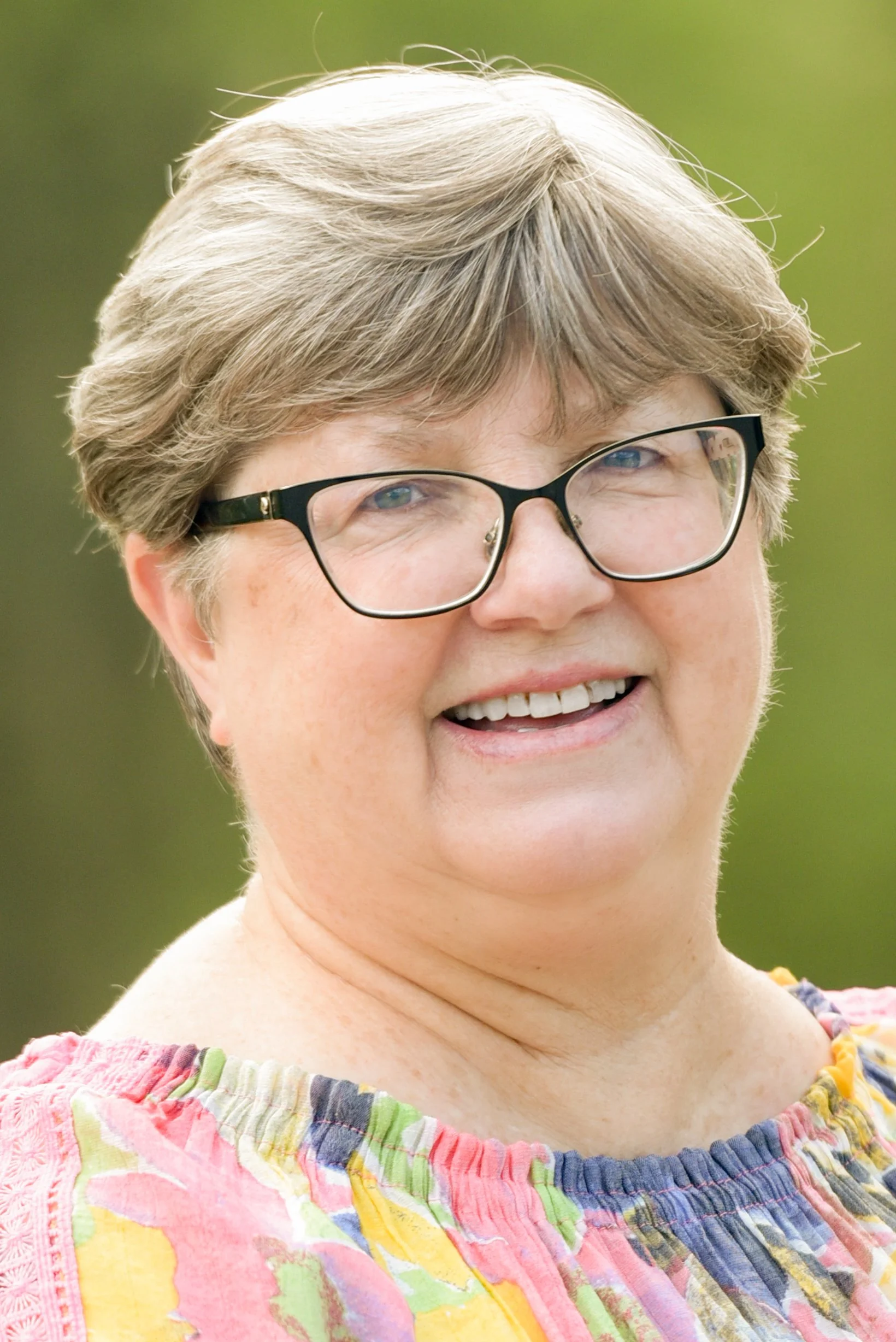 A smiling woman with short hair, glasses, and a colorful, patterned top outdoors with a blurred green background.