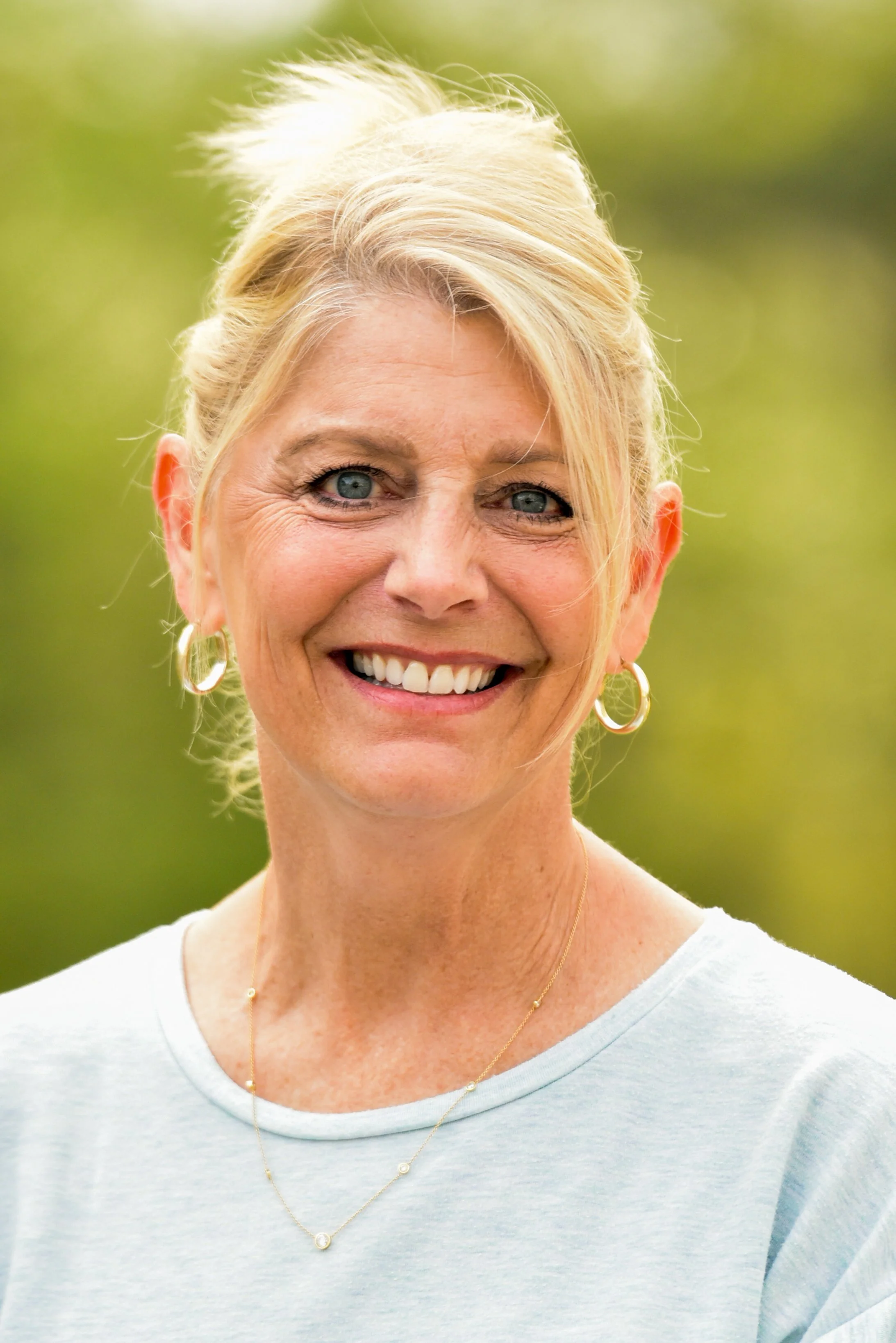 Close-up of a smiling woman with blonde hair, wearing earrings, a light gray T-shirt, and a delicate necklace, outdoors with a blurred green background.