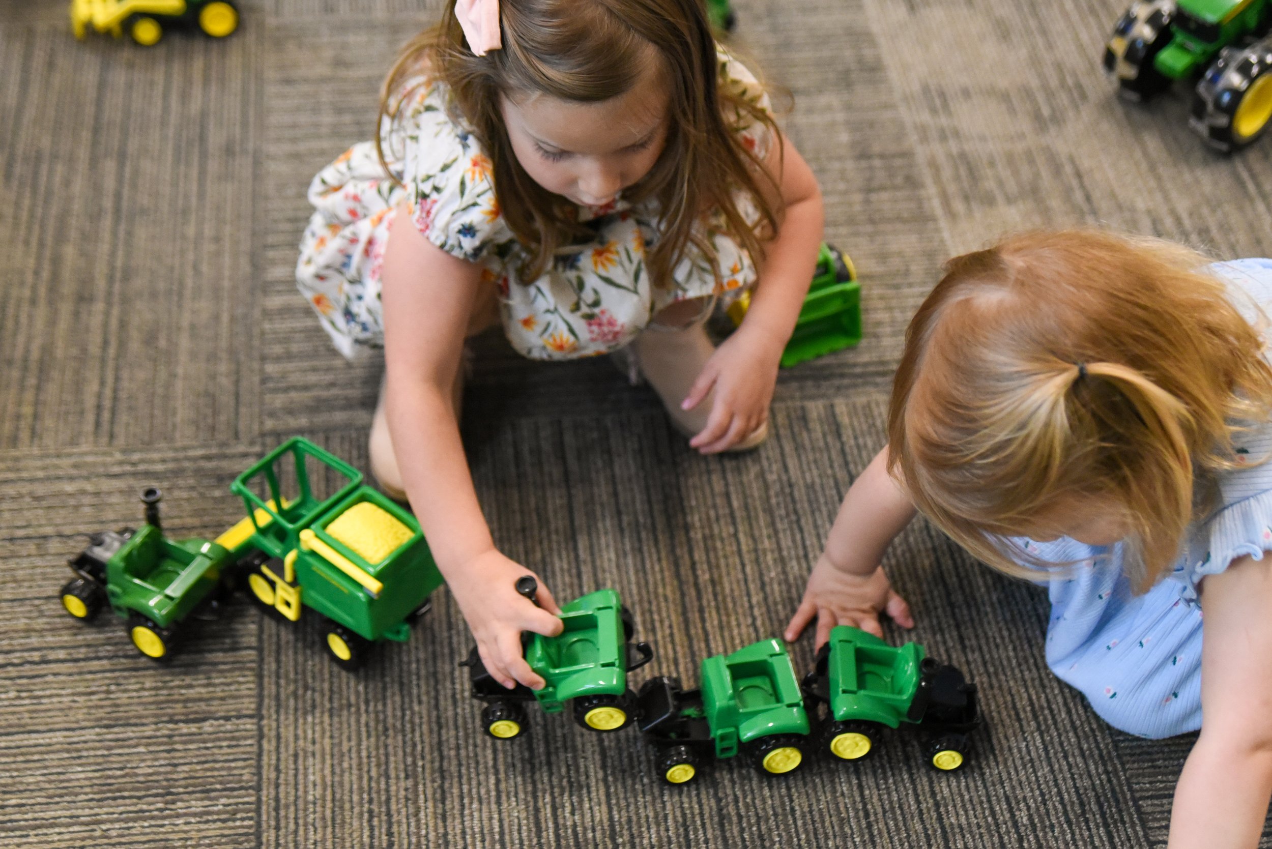 Two children playing with small green and black toy tractors on a carpeted floor.