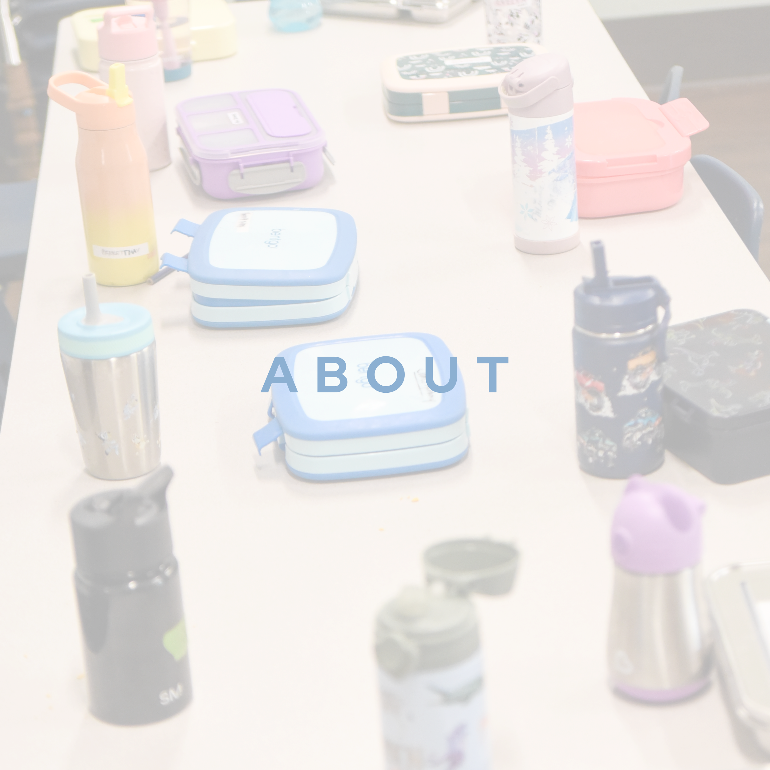Close-up of a table with various colorful personal items including water bottles, snack containers, and a pencil case, with the word 'ABOUT' visible on a paper in the foreground.