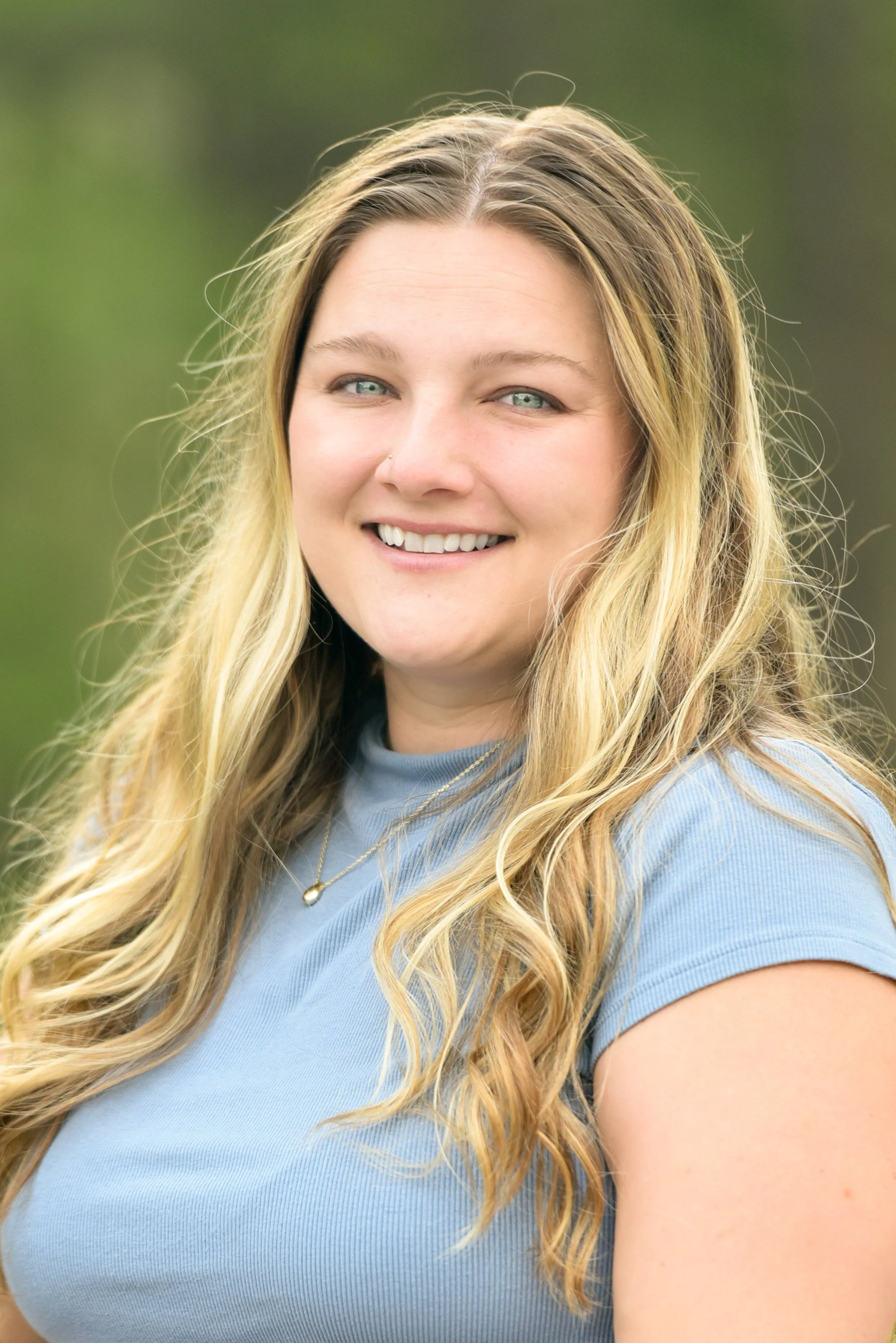 A young woman with long blonde hair, blue eyes, smiling outdoors with a blurred green background. She wears a light blue short-sleeved top, a delicate necklace, and a gold necklace.