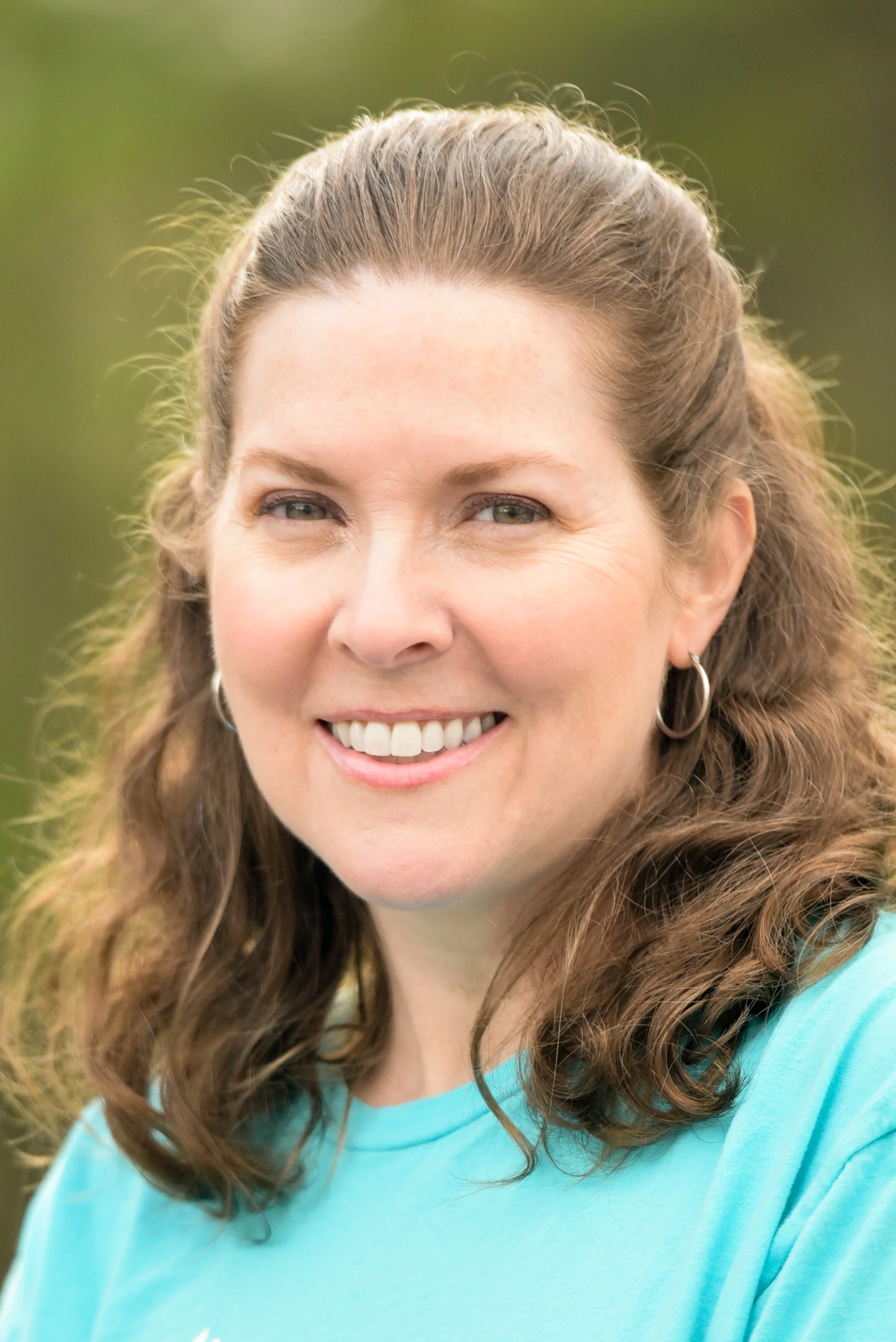 Close-up of a smiling woman with curly brown hair wearing a light blue shirt and hoop earrings, outdoors with green background.