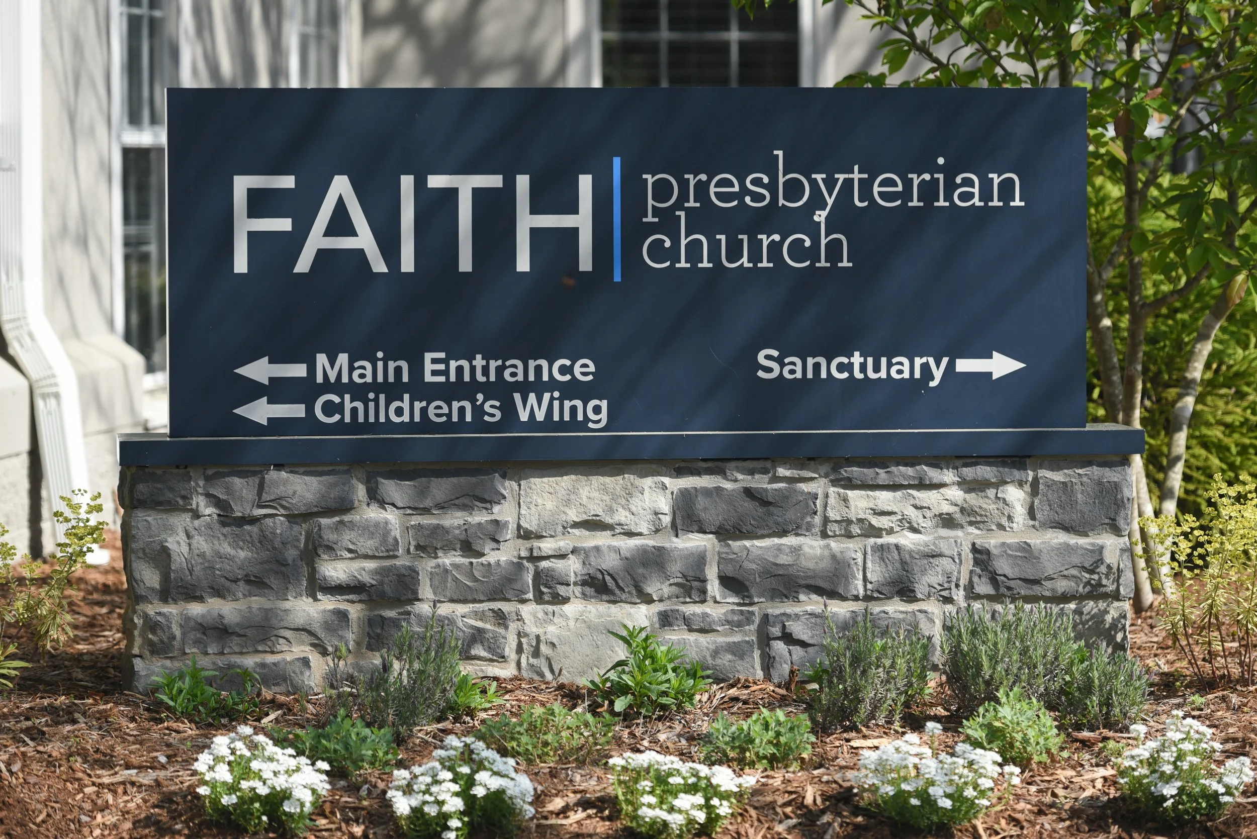 Stone sign for Faith Presbyterian Church with directions for main entrance, children's wing, and sanctuary, surrounded by landscaped plants and trees.