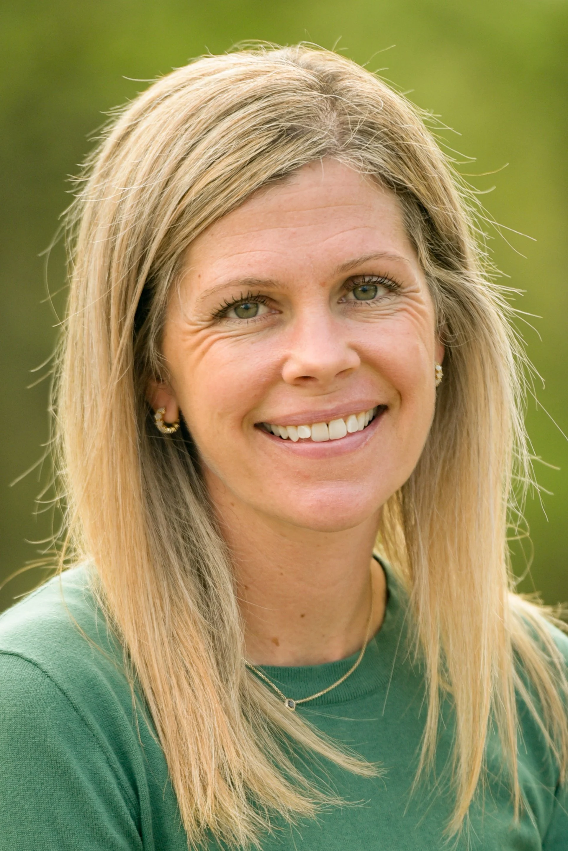 A woman with blonde hair wearing a green shirt and gold jewelry, smiling outdoors with a blurred green background.