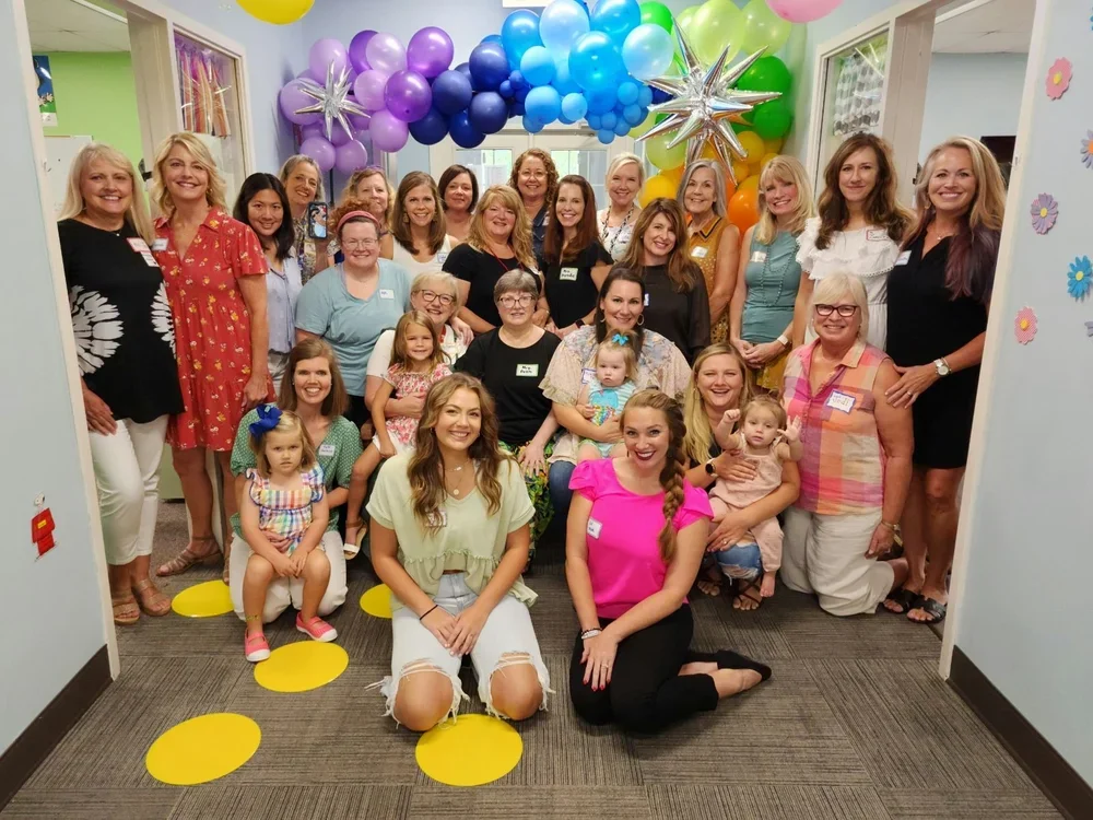 Group of women and children at a celebration indoors, decorated with colorful balloons and paper flowers.