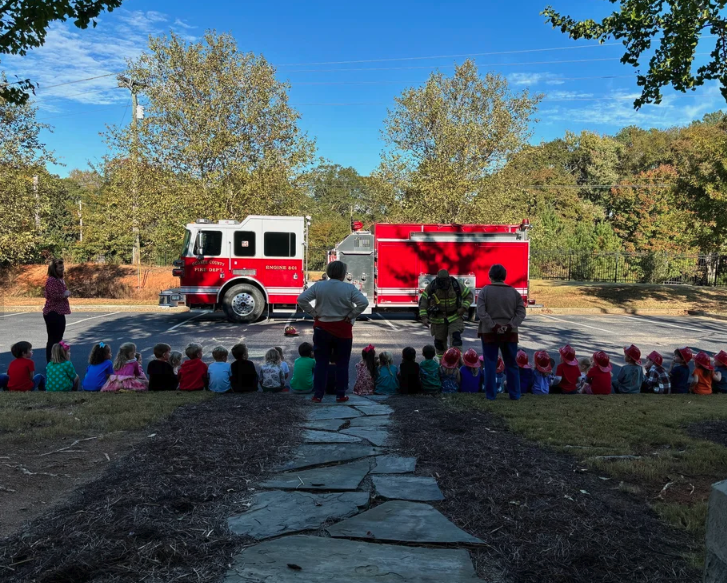 Children sitting on a grassy area watching a fire truck showcase surrounded by adults, with trees and a blue sky in the background.