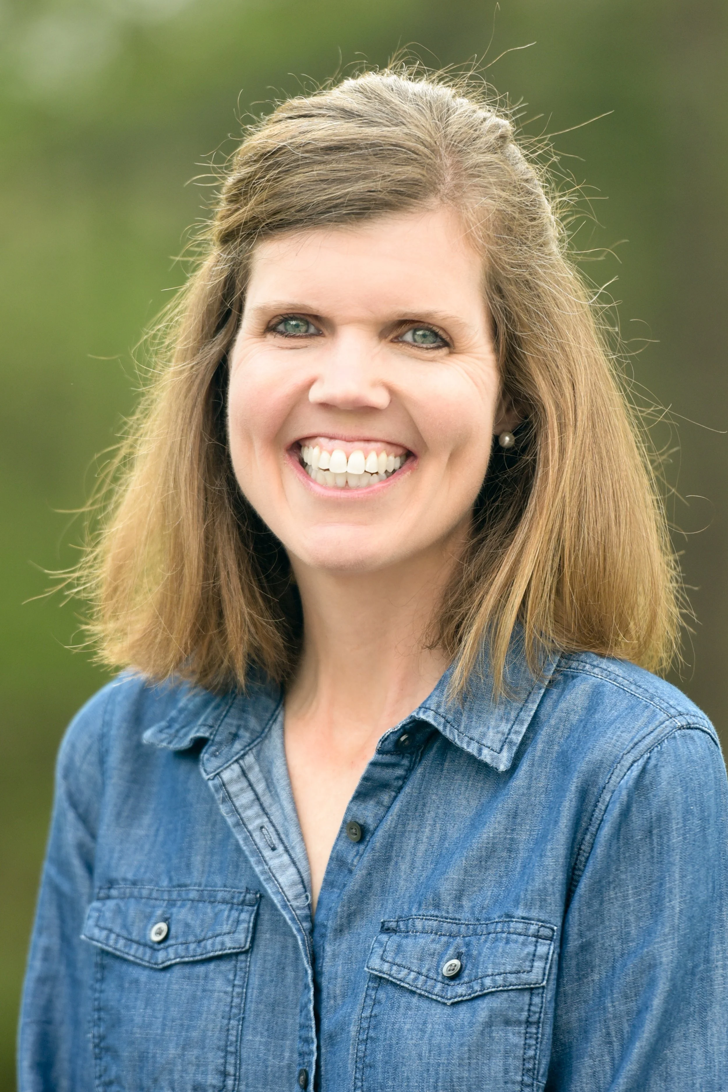 A woman smiling outdoors with green blurred background, wearing a denim shirt and pearl earring.