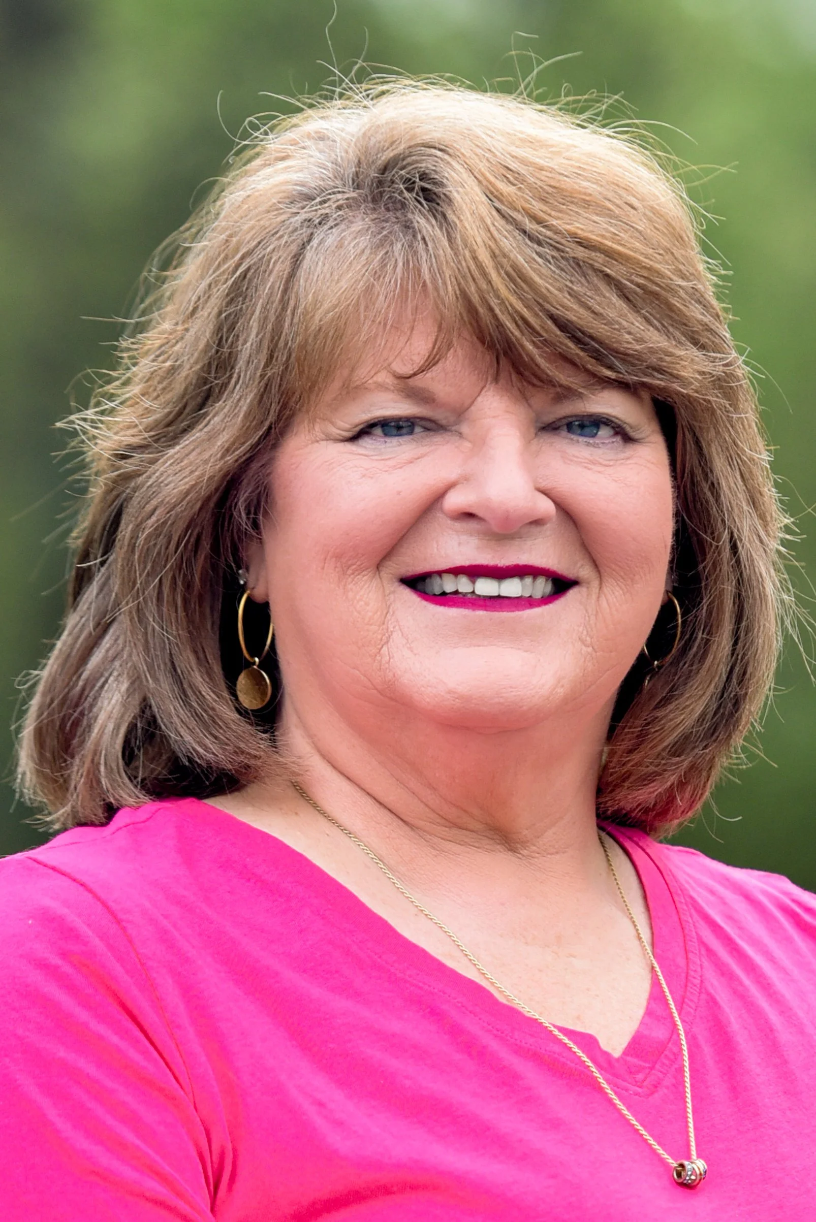 A smiling woman with light brown hair, wearing a pink top, gold earrings, and a gold necklace with a pink pendant. She has a background of blurred green outdoor scenery.