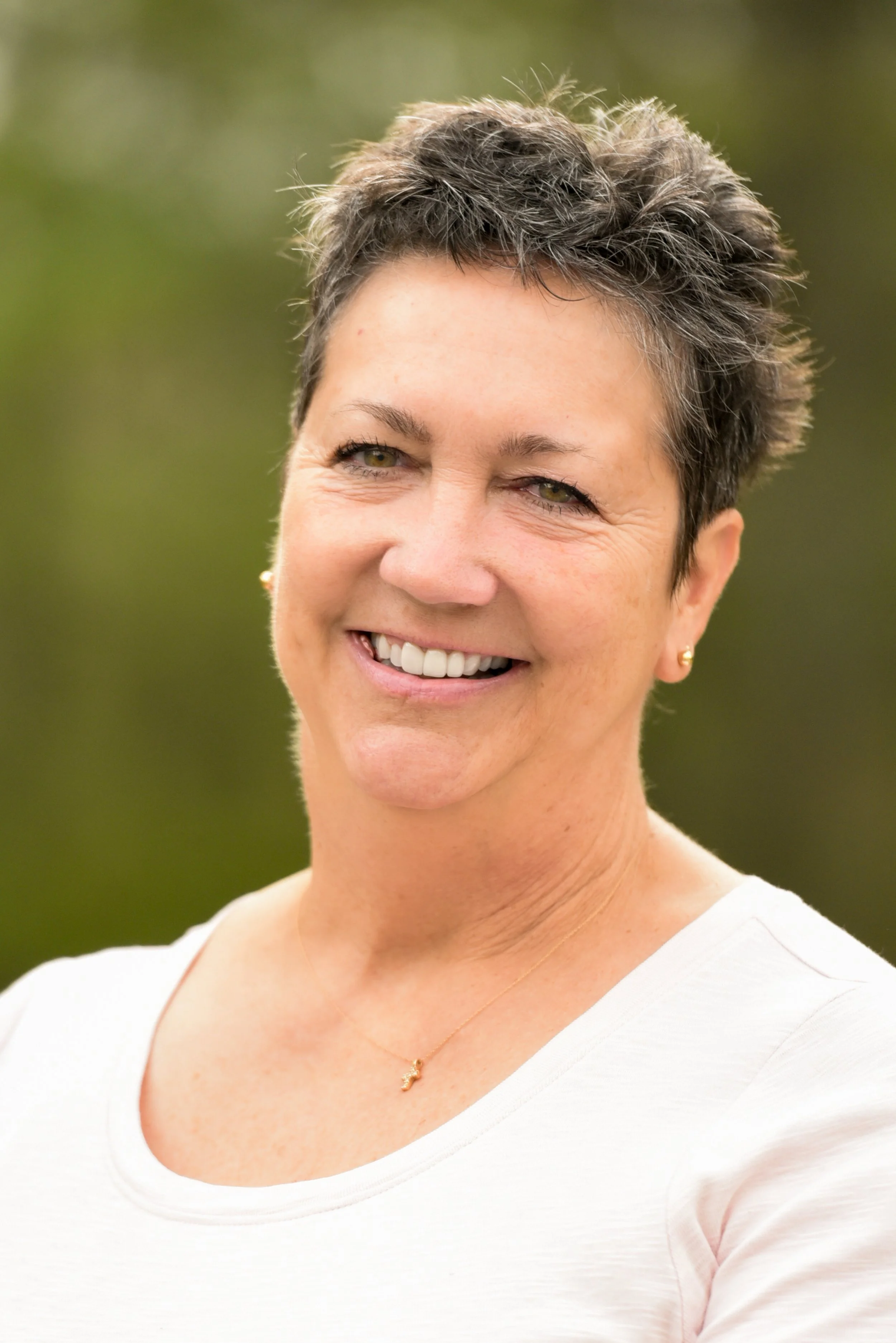 A woman with short, tousled gray and dark hair smiling outdoors with a blurred green background, wearing a white top, gold earrings, and a delicate gold necklace.