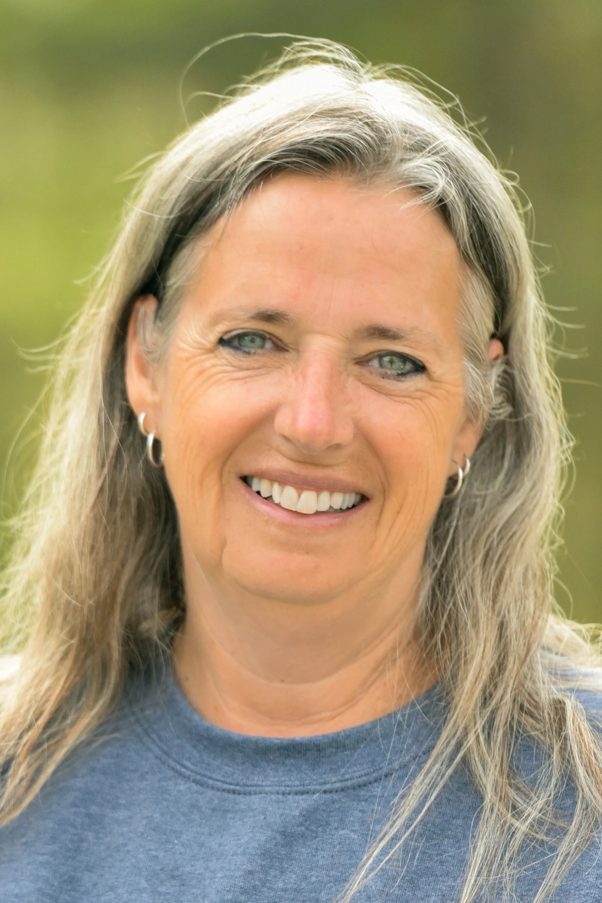 Close-up of a smiling woman wearing a blue shirt, outdoors with blurred green background.