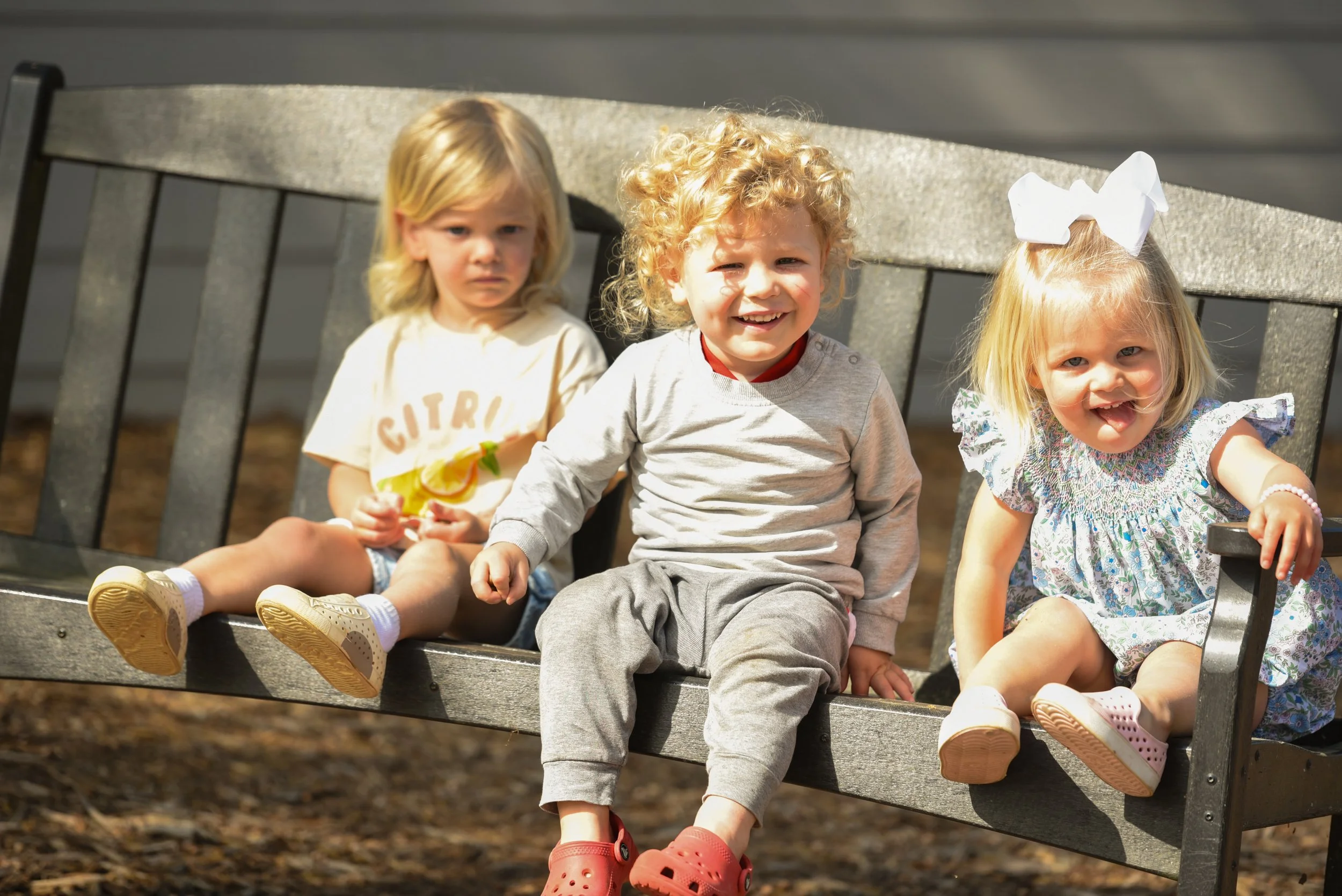 Three young children sitting on a park bench outdoors, with two girls and one boy. The girl on the left has blonde hair and is wearing a yellow T-shirt, the boy in the middle has curly blonde hair and is smiling, wearing a gray shirt and pants, and t