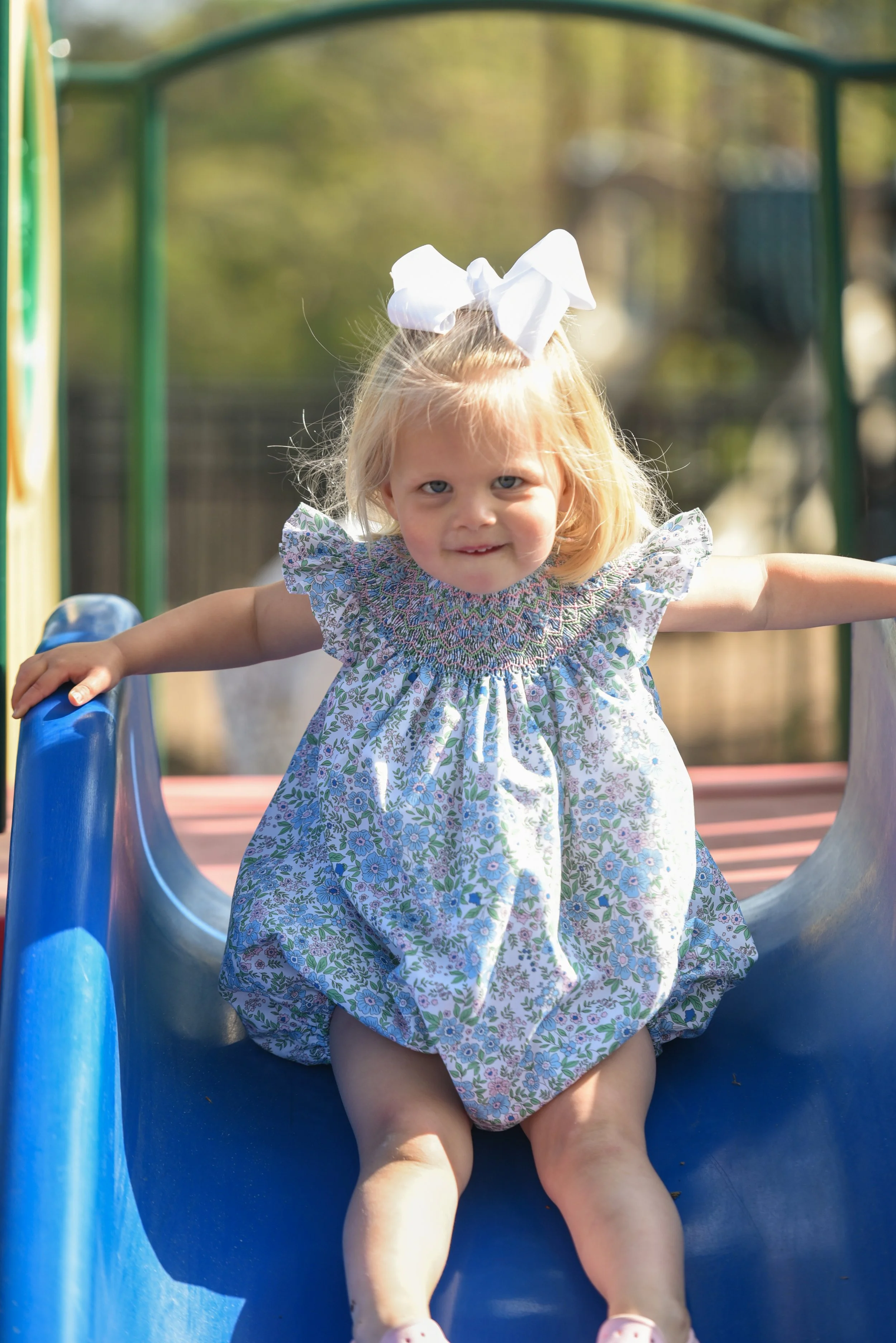 A young girl with blonde hair tied with a large white bow, wearing a floral dress, is on a blue playground slide outdoors. She has her arms outstretched, and a slight smile on her face, with sunlight illuminating her.