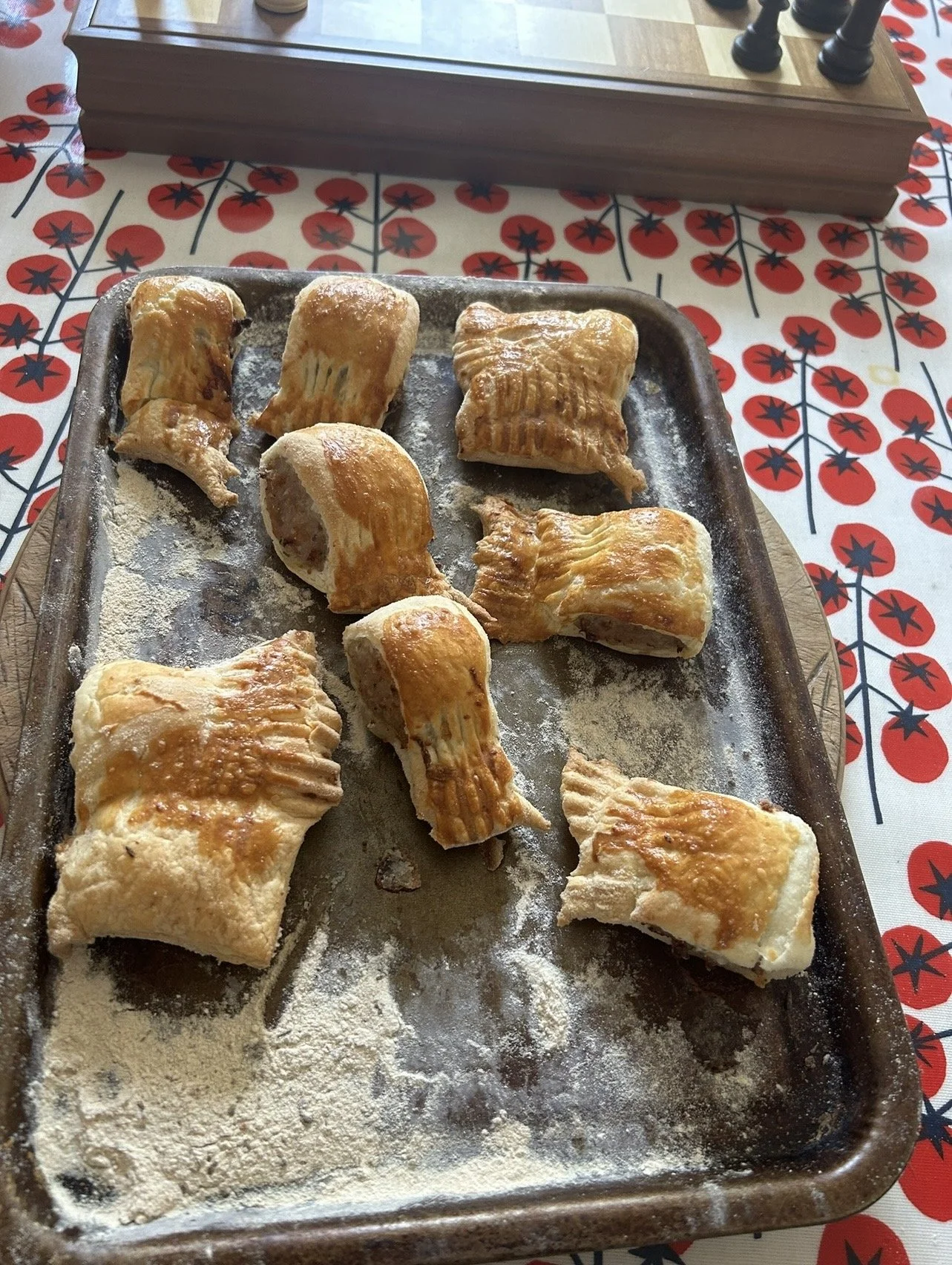 Baked sausage rolls on a baking sheet with flour dusting, placed on a festive tablecloth with red berries design, and a wooden box with chess pieces in the background.