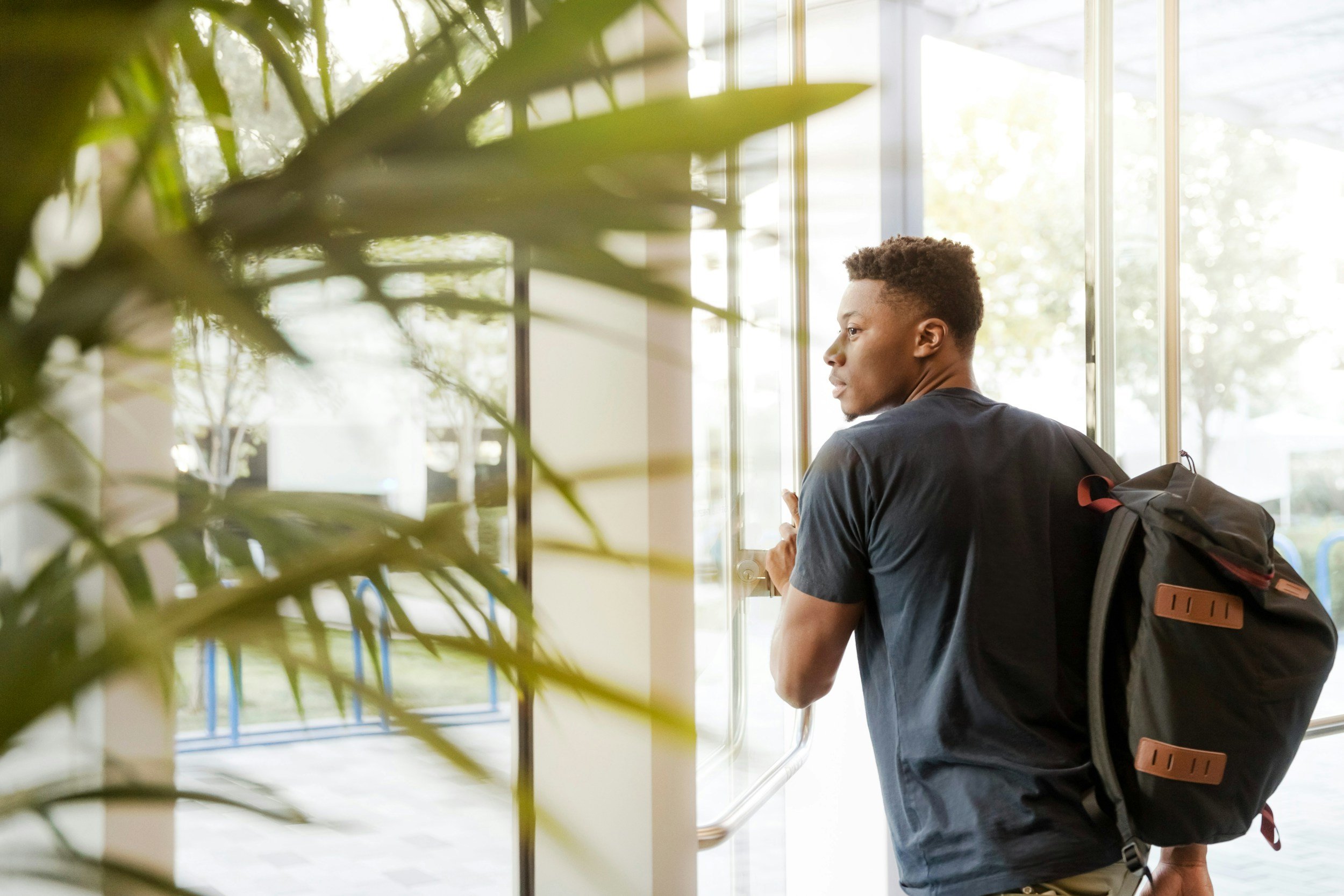 A young man with short hair, wearing a dark t-shirt and carrying a backpack, is standing by a glass door, looking outside in a bright, sunlit area with trees visible through the window.