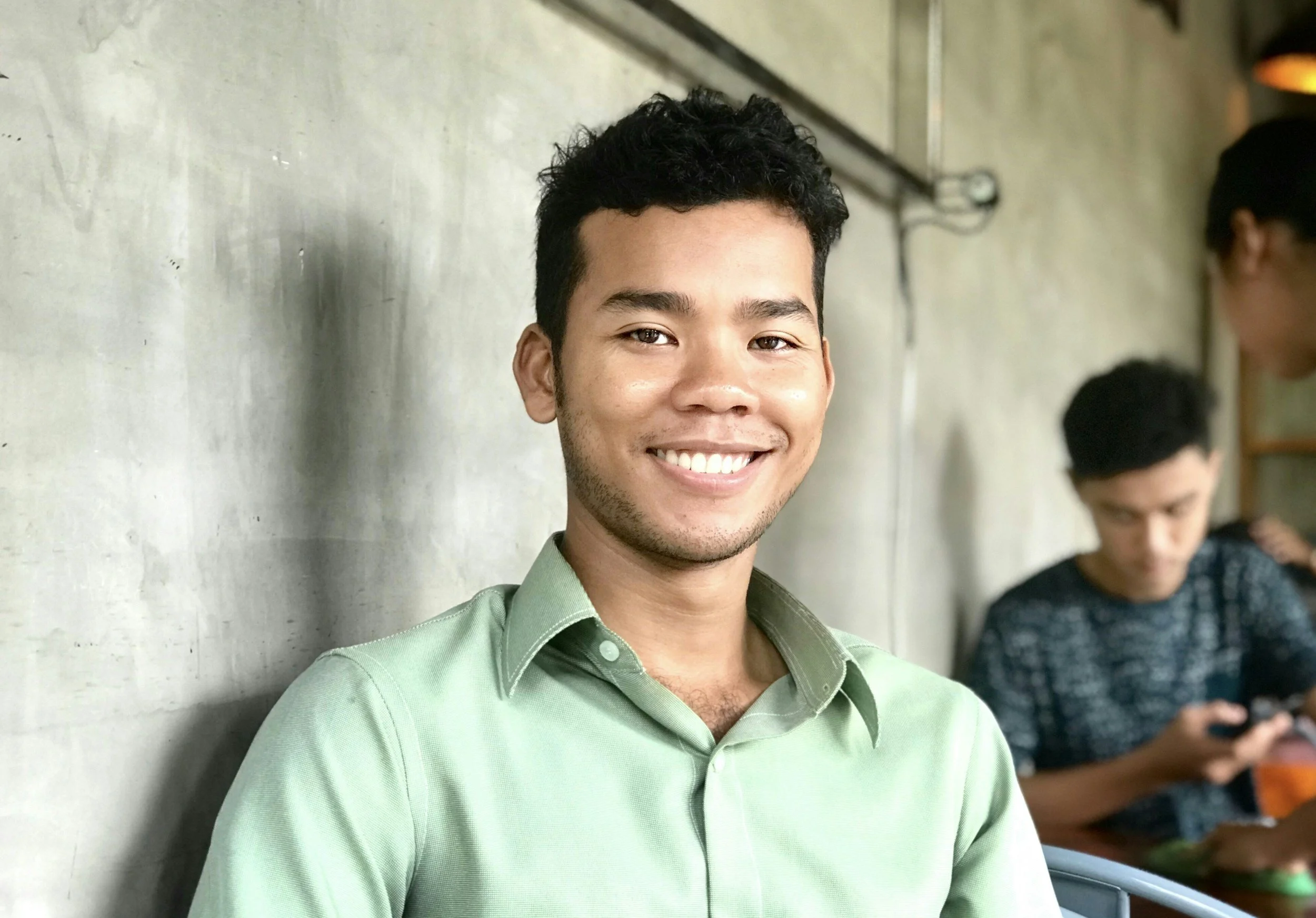 A young man with curly black hair and a light green button-up shirt smiling and leaning against a concrete wall. In the background, two other young men are engaged with their phones.