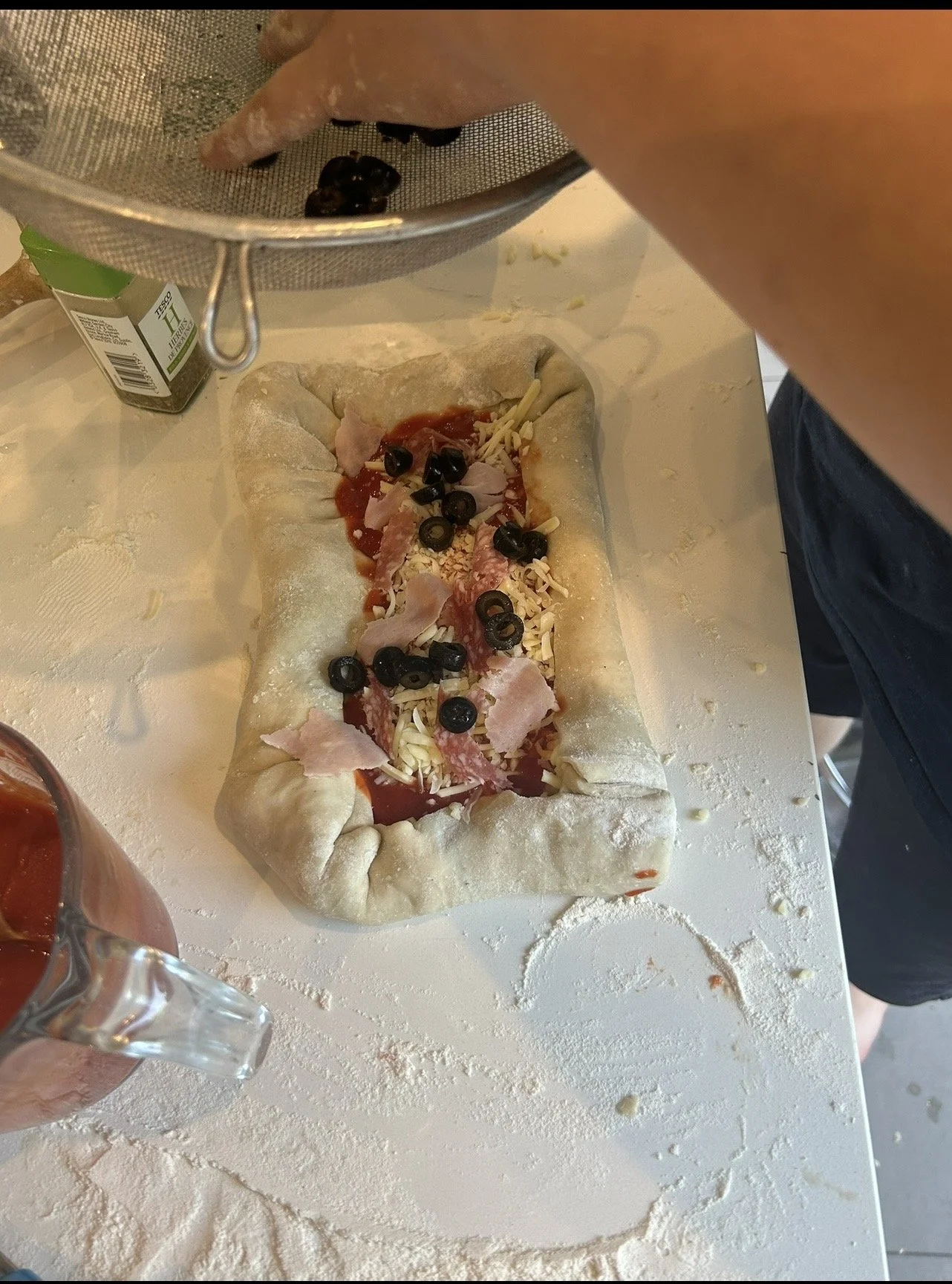 Person preparing a rectangular pizza with tomato sauce, shredded cheese, sliced black olives, ham, and bacon on a floured surface.