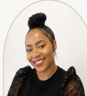 Smiling woman with dark hair styled in a bun, wearing a black top with sheer, dotted sleeves, in front of a white background.