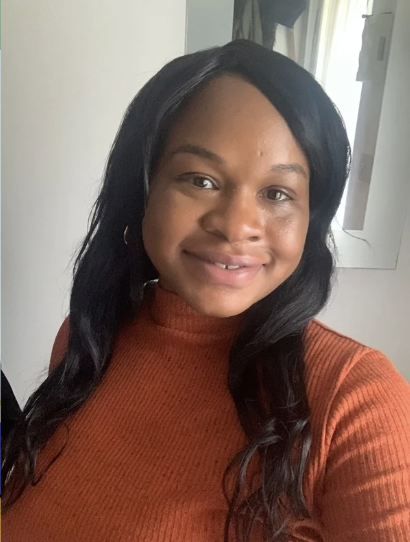 A young woman with long black hair, wearing a burnt orange turtleneck sweater, smiling at the camera inside a room.