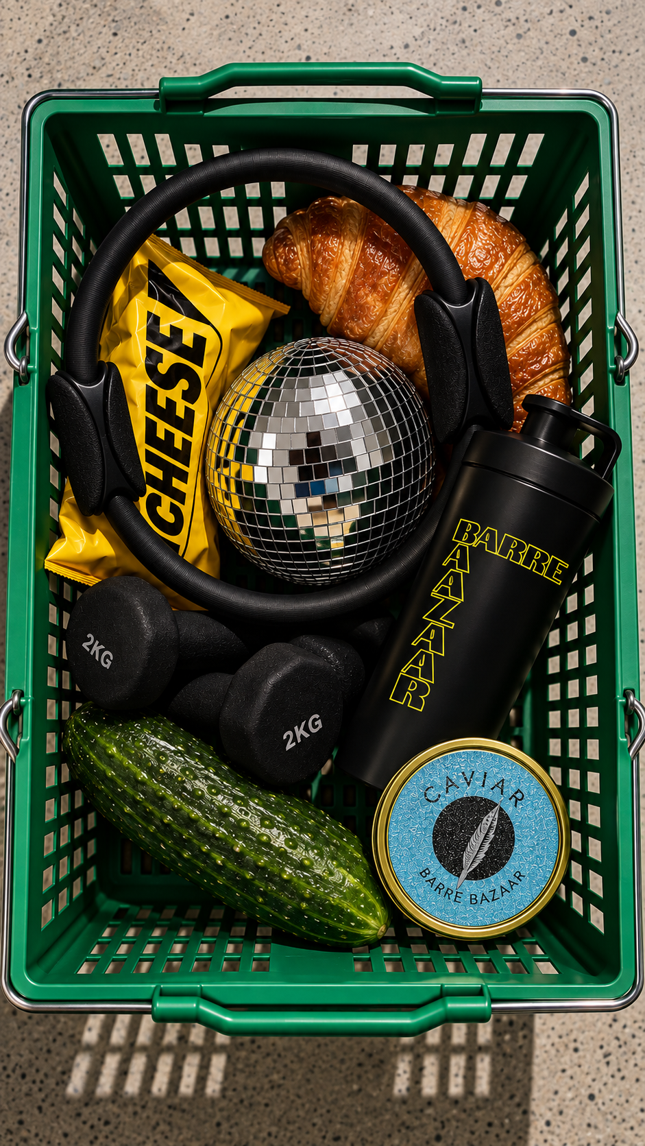 Green shopping basket containing a croissant, a yellow package labeled 'CIEESE', a disco ball, a black water bottle labeled 'BARRE BAZAAR', two 2kg dumbbells, a cucumber, and a tin of caviar, on a speckled floor.