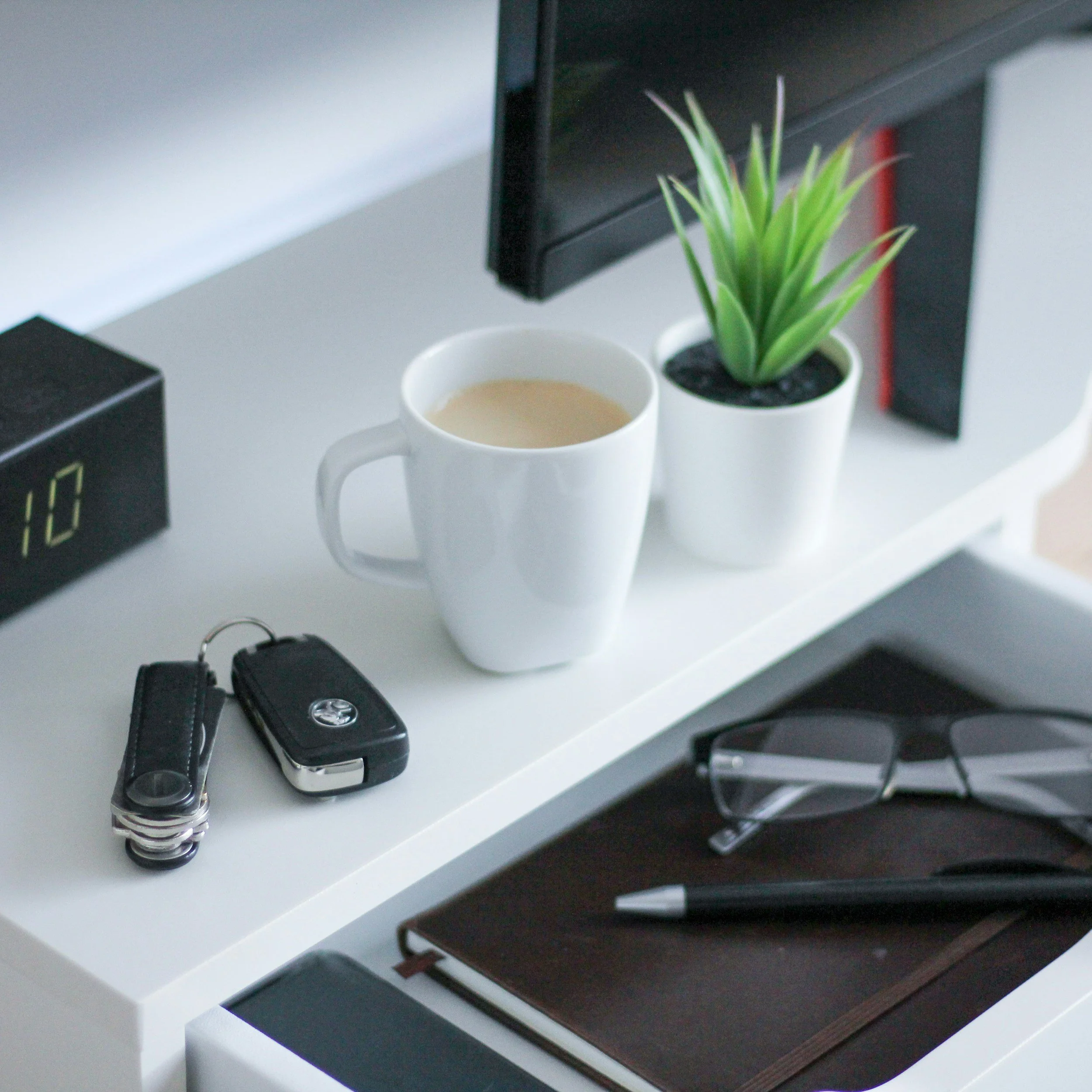 A white shelf with a white mug filled with coffee, a small green potted plant, a black electronic device, and black framed books or boxes.