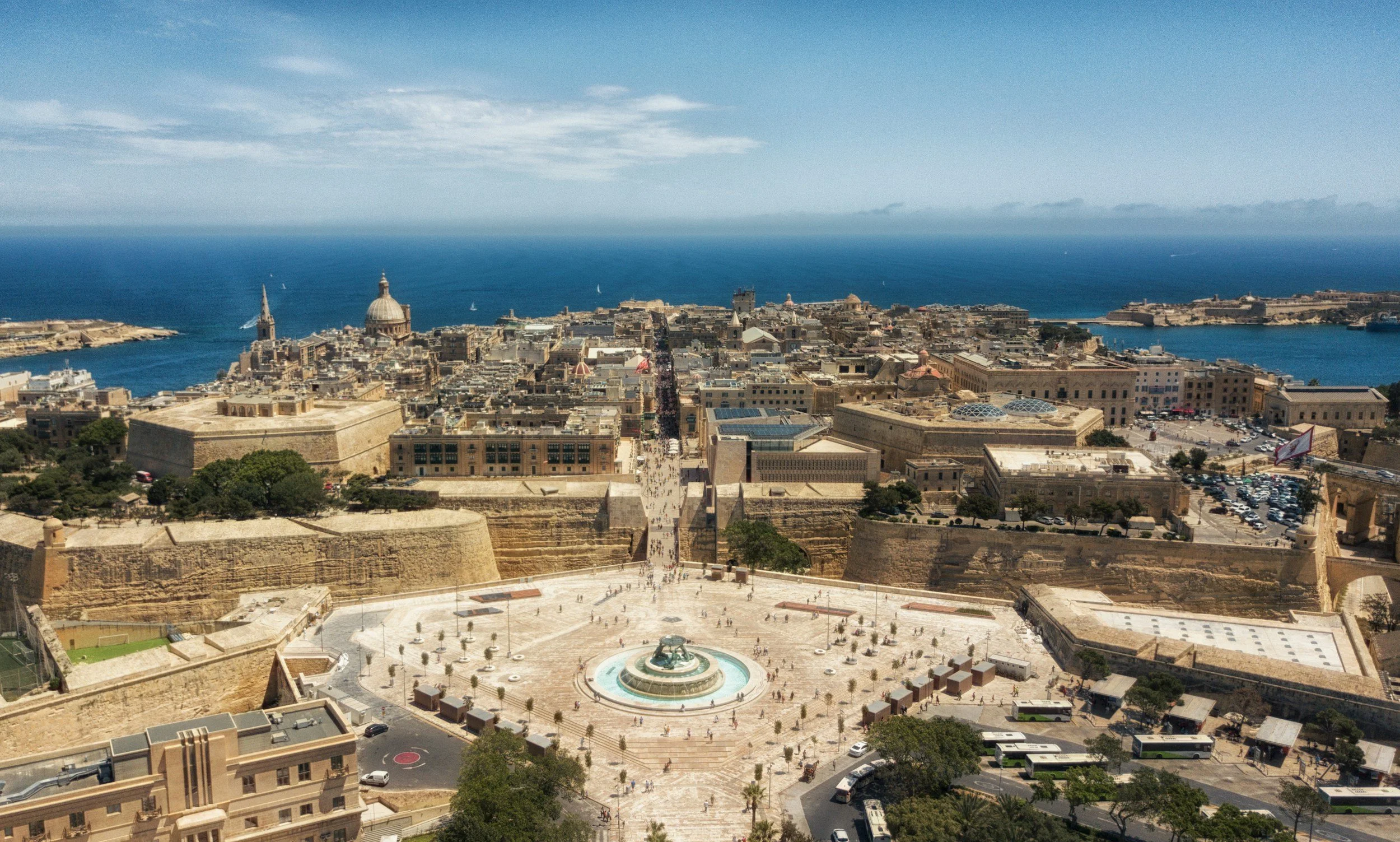 A panoramic view of Valletta, Malta, featuring historic buildings, a large open square with a fountain, and the Mediterranean Sea in the background.