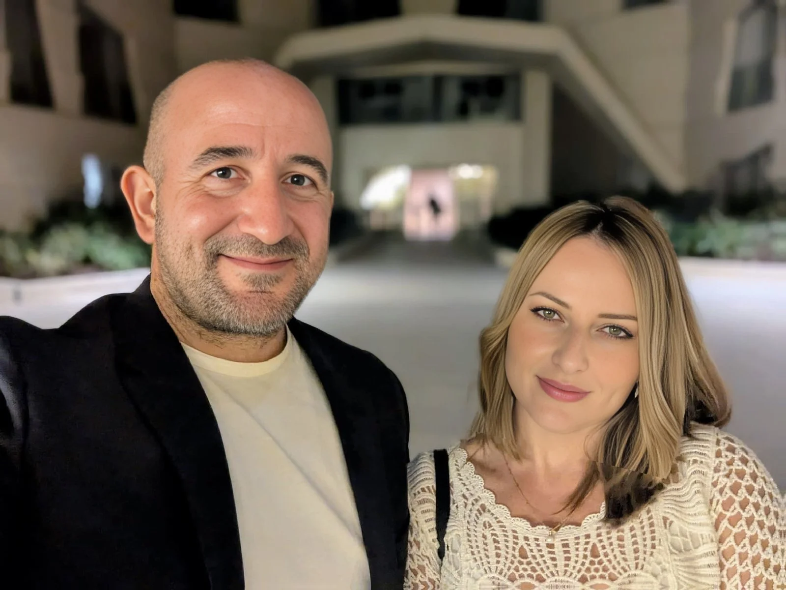 A man and woman taking a selfie outdoors at night in front of a modern building with large windows and plants.