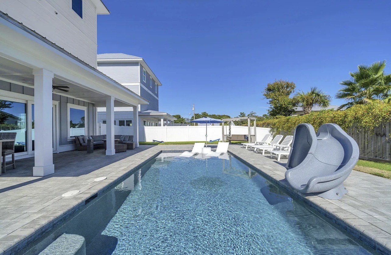 Empty backyard swimming pool with lounge chairs and umbrellas, patio furniture, and a white fence, in a sunny residential area with clear blue sky.
