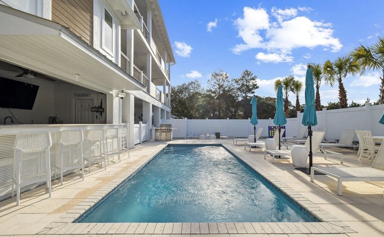 Empty swimming pool area with lounge chairs and blue umbrellas, white fence, in a sunny backyard with palm trees and a blue sky.