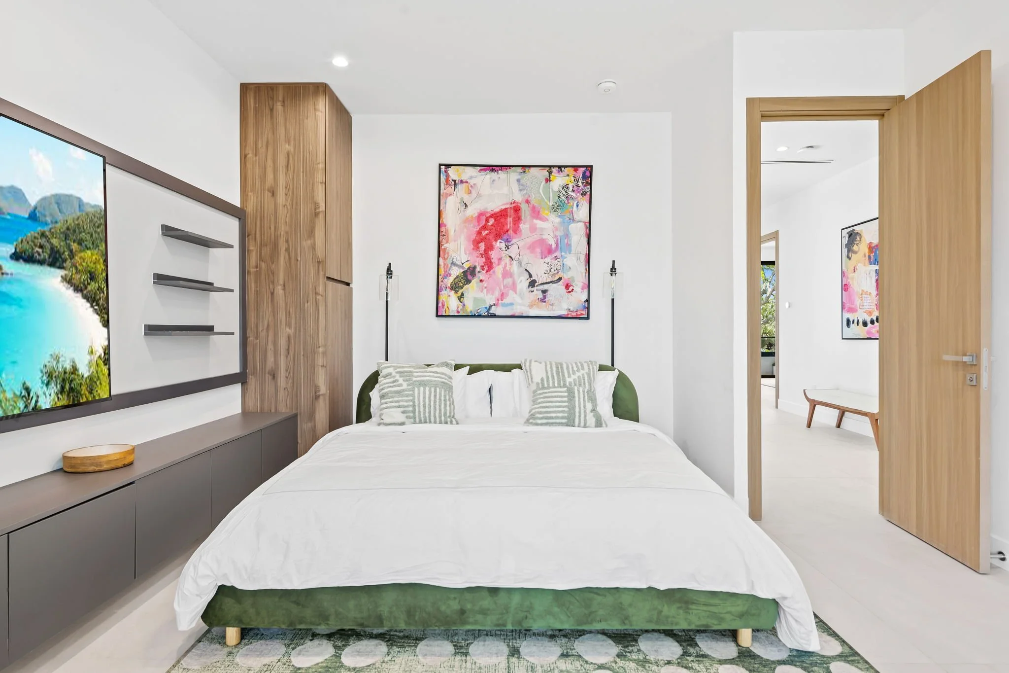 Modern bedroom with a white bed, green upholstered frame, patterned pillows, wall art, wooden accents, and a doorway leading to another room with artwork and a window.